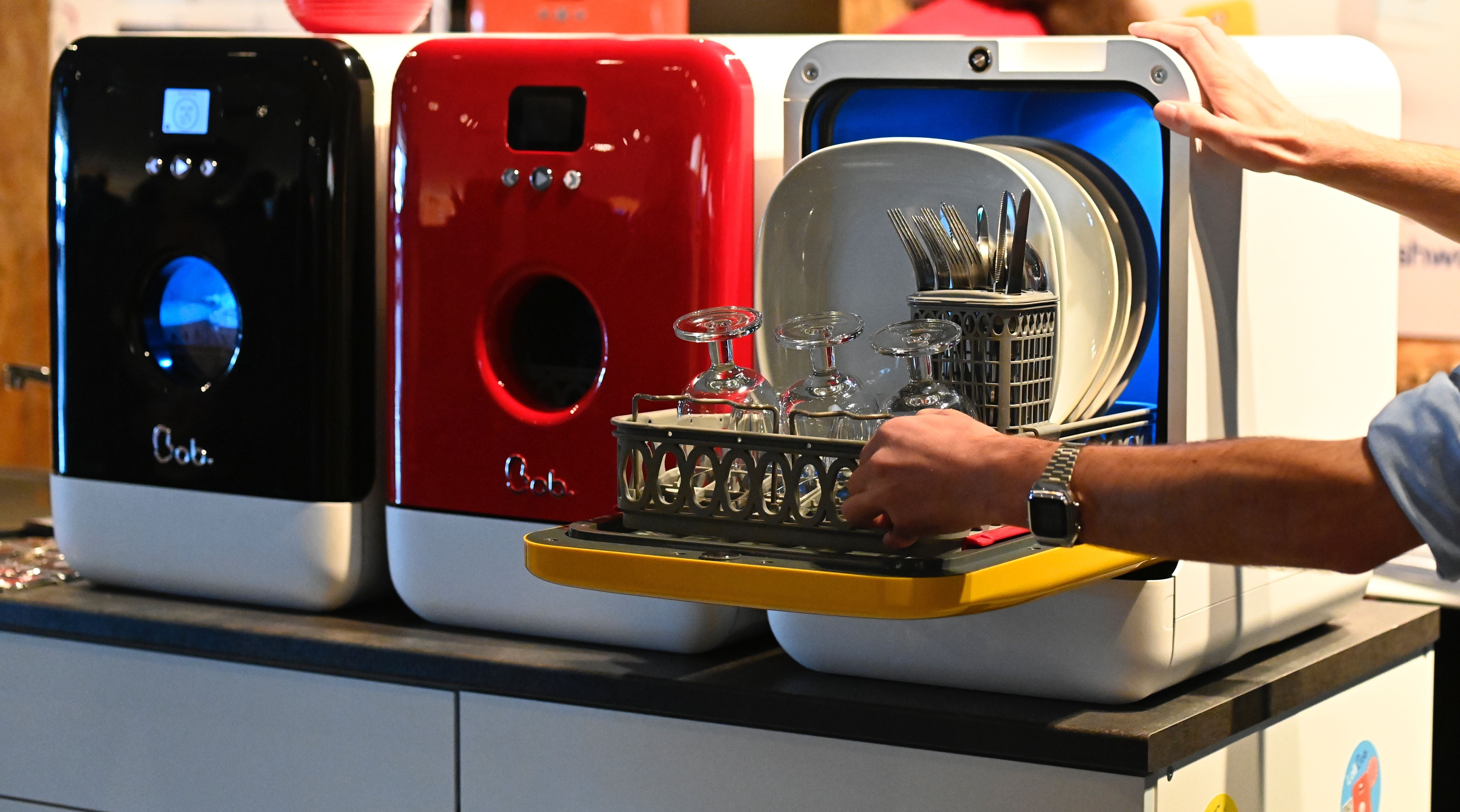 Person removing a rack of cleanable  dishes and glasses from a compact countertop dishwasher