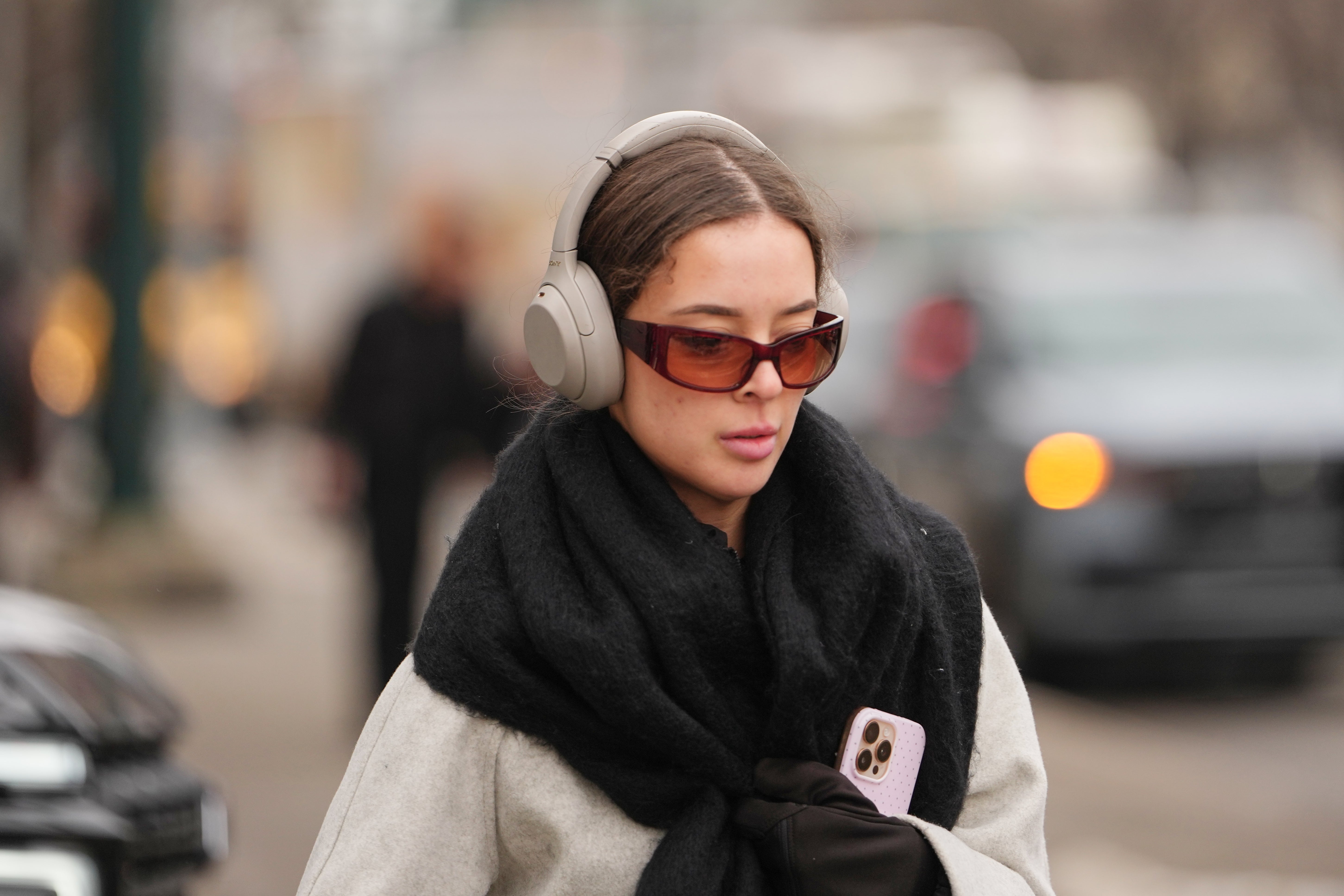Person walking extracurricular  wearing ample  headphones, sunglasses, and a scarf, holding a smartphone
