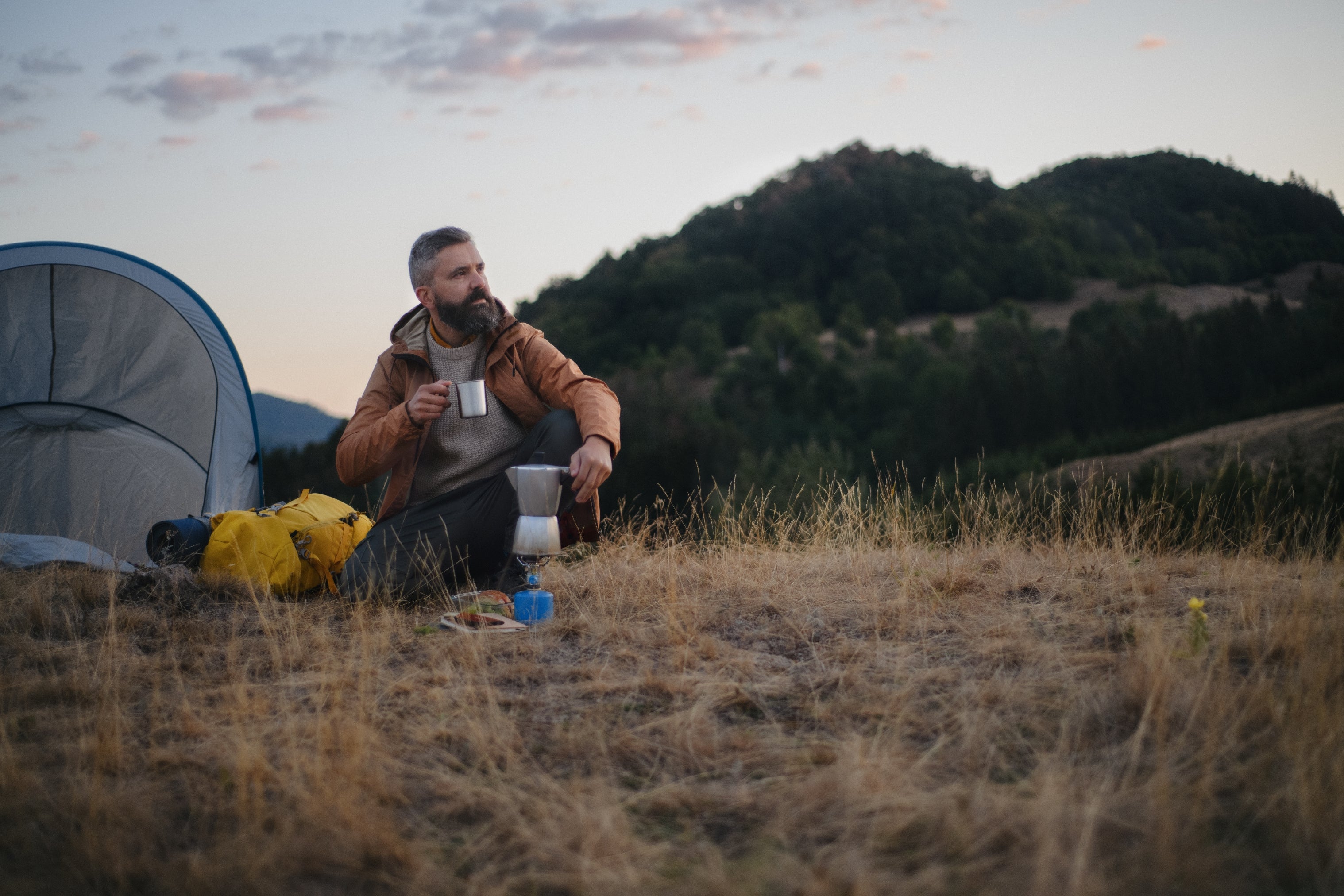 Man sitting by a structure   successful  a field, holding a mug, gazing astatine  the mountains. Camping cogwheel  lies nearby, suggesting a serene outdoor setting