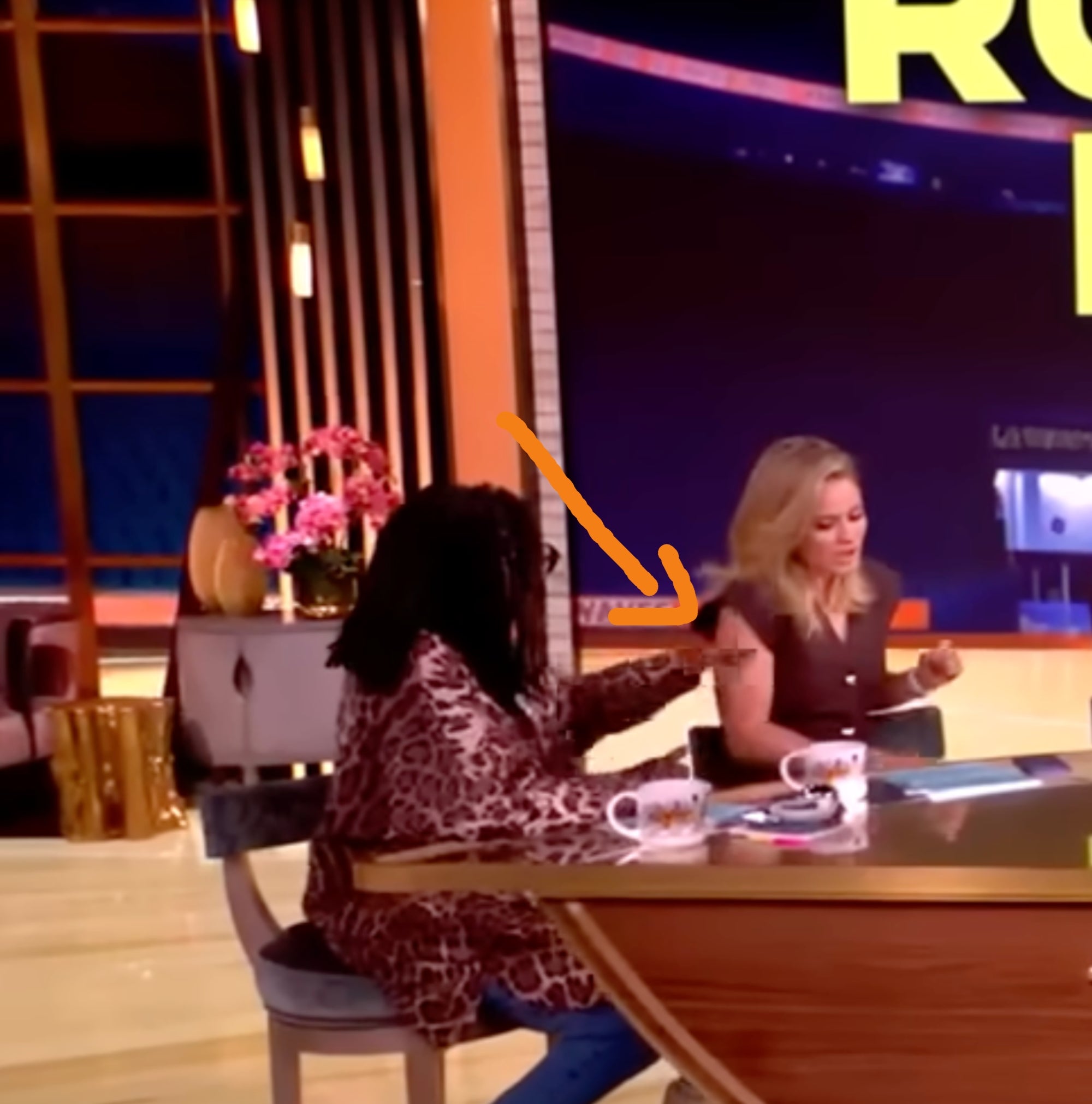 Two radical   seated astatine  a TV workplace  table  are engaged successful  a conversation, with cameras and workplace  lights disposable   successful  the background