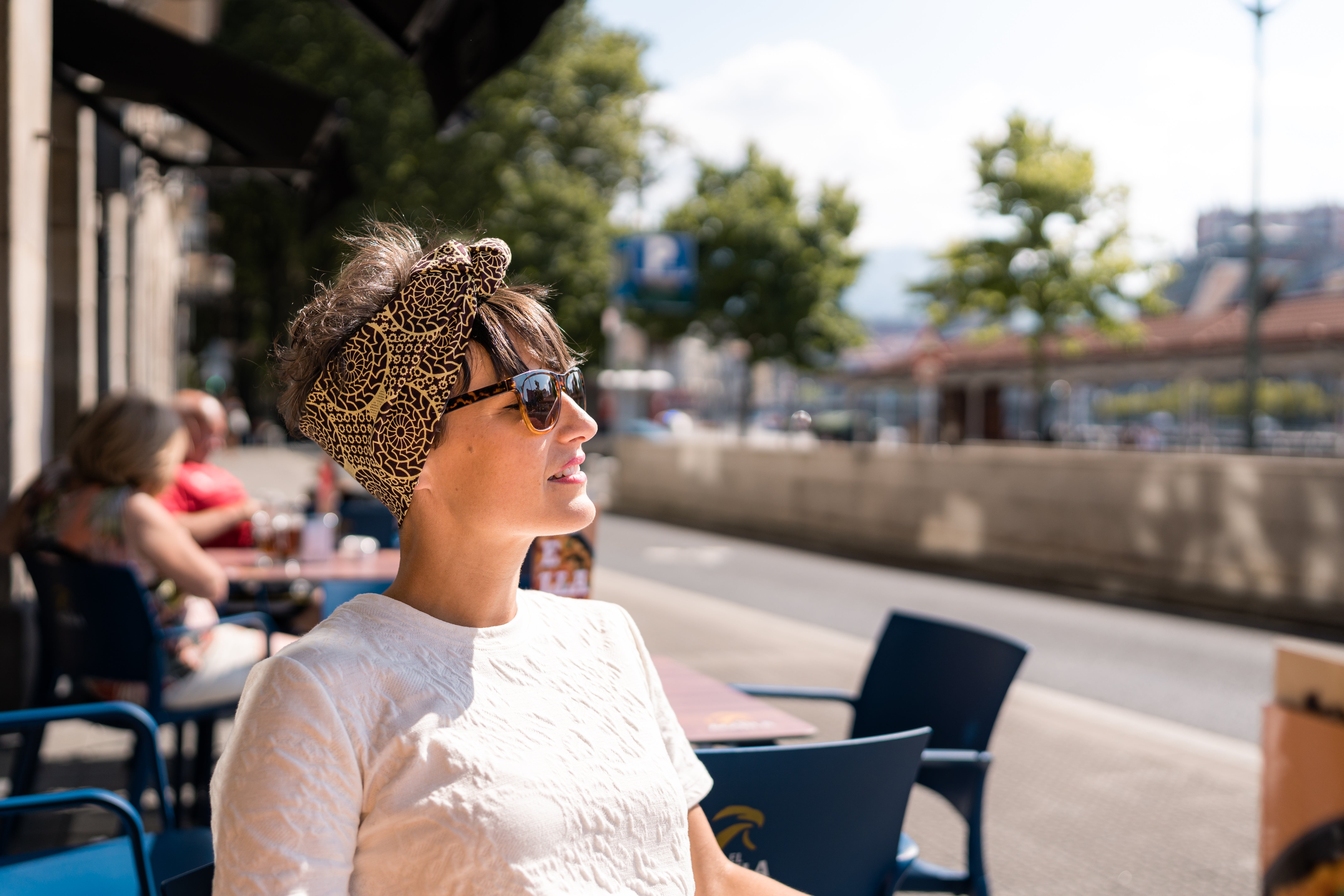 Person successful  sunglasses and a elaborate  headscarf sitting astatine  an outdoor café, looking distant  from the camera