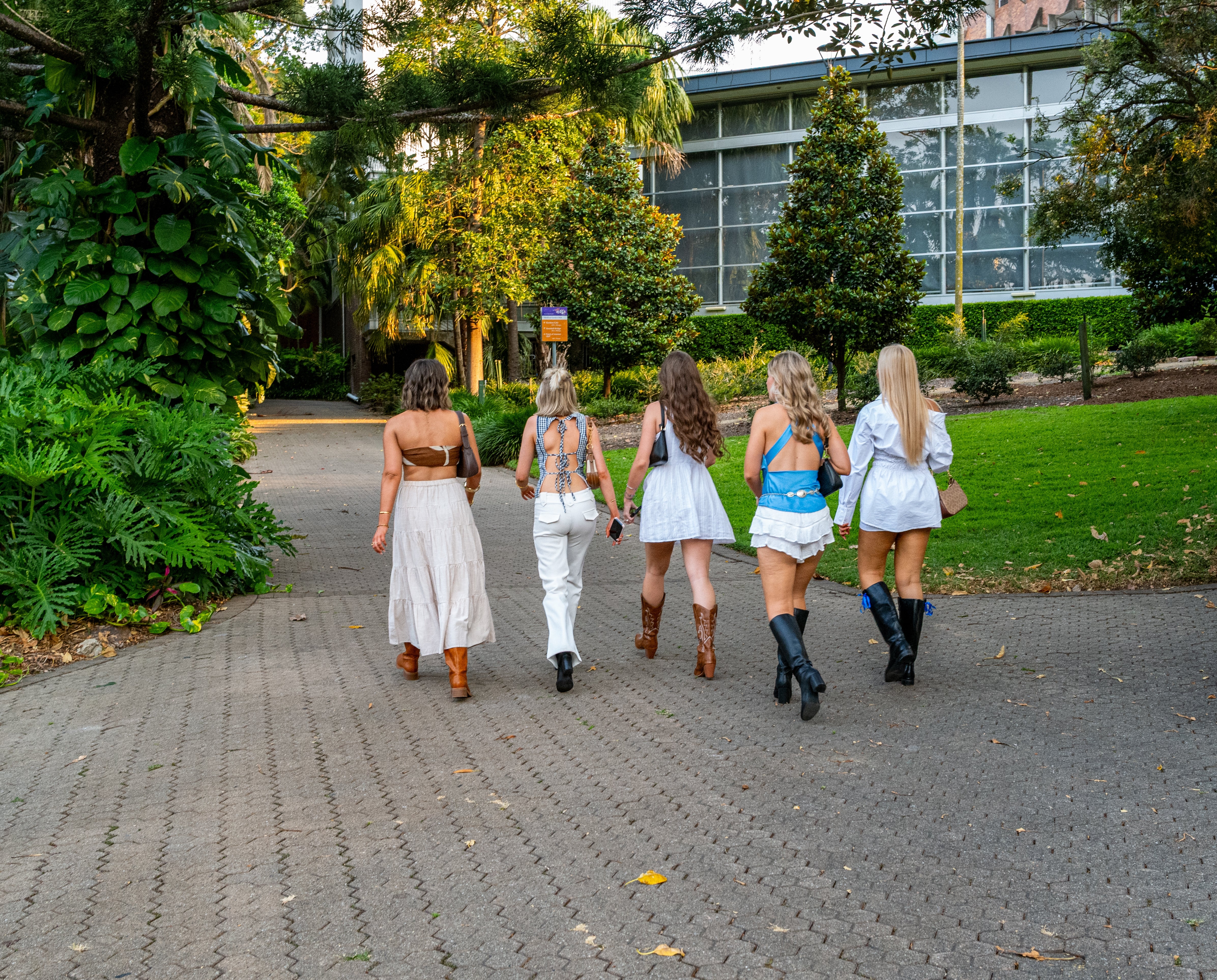 Five women from down  successful  casual summertime  outfits and boots, walking connected  a paved way  lined with greenery