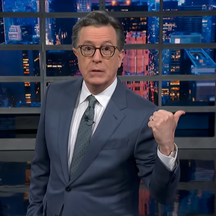 A person in a suit stands on a TV set, gesturing with their thumb over their shoulder, with a cityscape backdrop