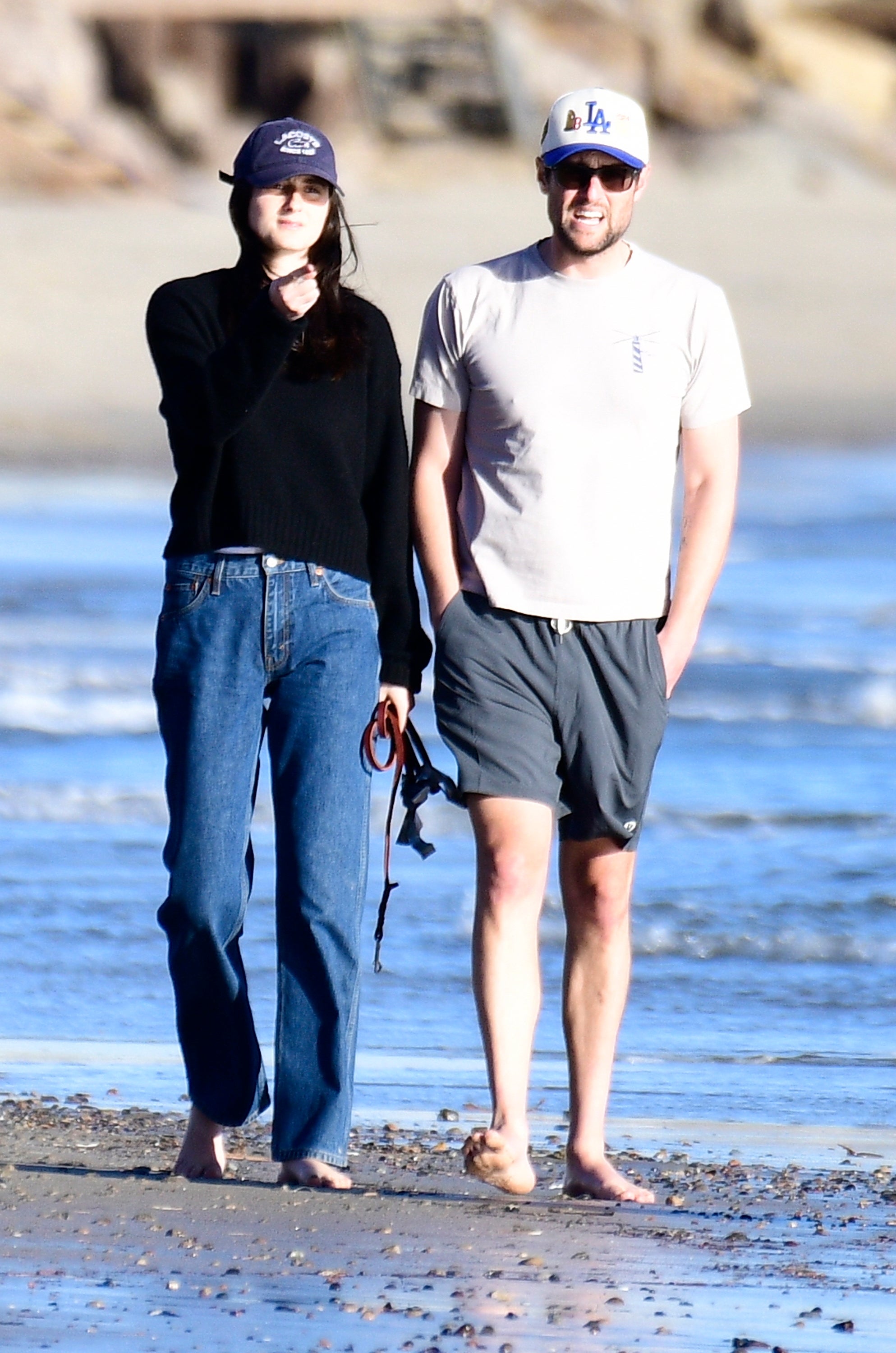 Two radical   walking connected  the beach. One wears a achromatic  sweater and jeans, the different   a achromatic  t-shirt and shorts. Both deterioration  shot   caps