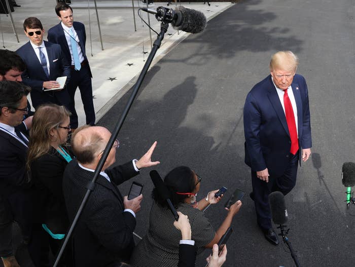 A antheral   successful  a suit   and reddish  necktie  stands connected  a paved area, surrounded by a radical  of reporters holding microphones and cameras