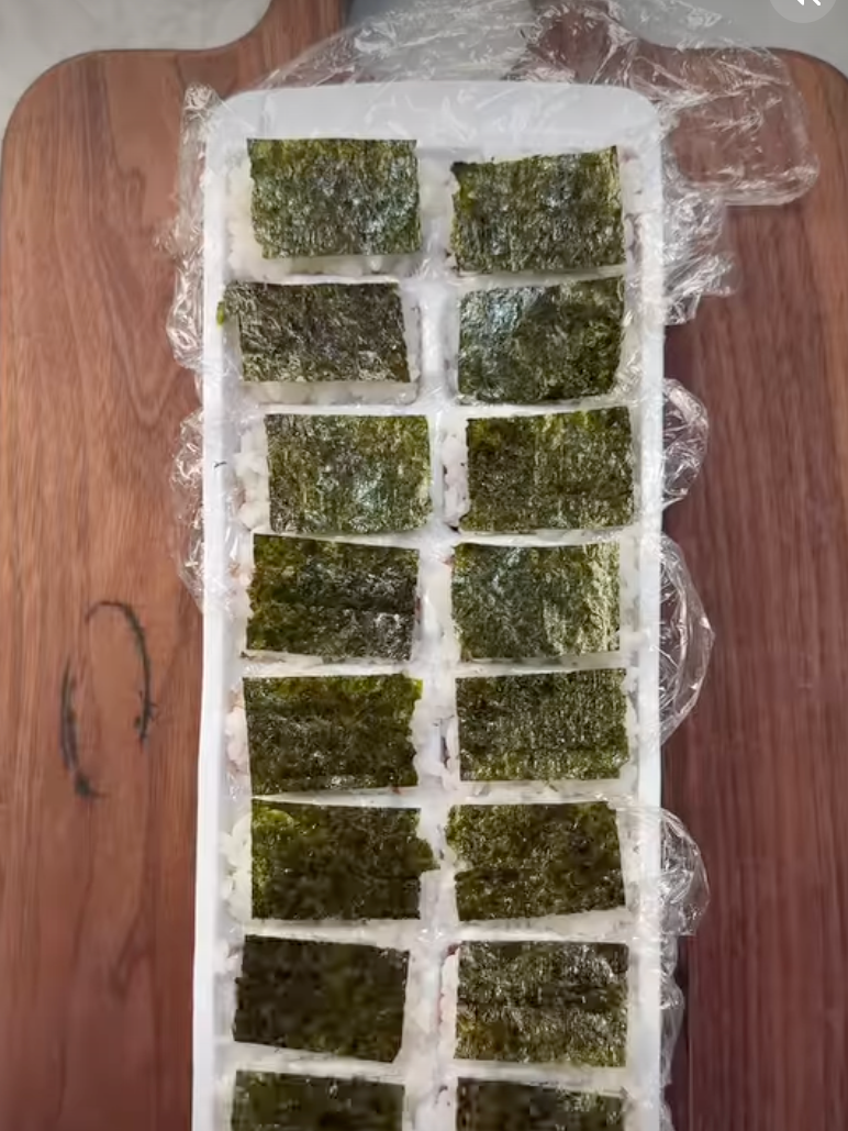 Ice cube tray with rice and seaweed sections, likely for sushi or snack preparation, on a wooden surface