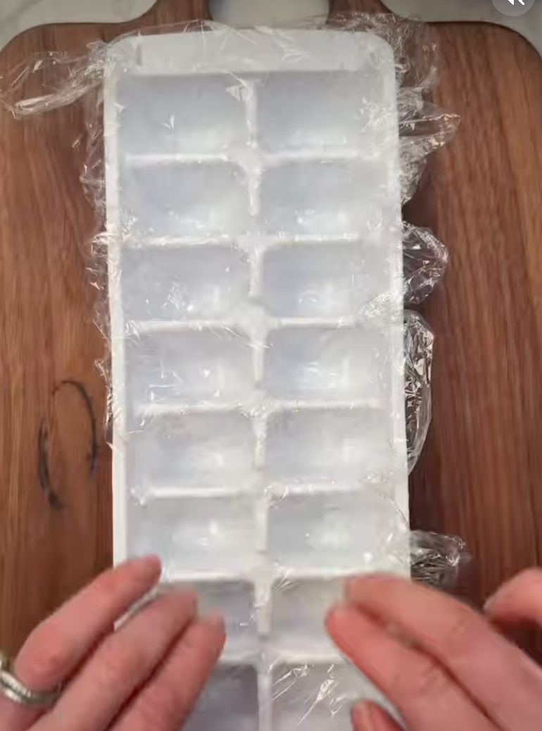 Hands wrap a plastic-covered ice cube tray on a wooden surface