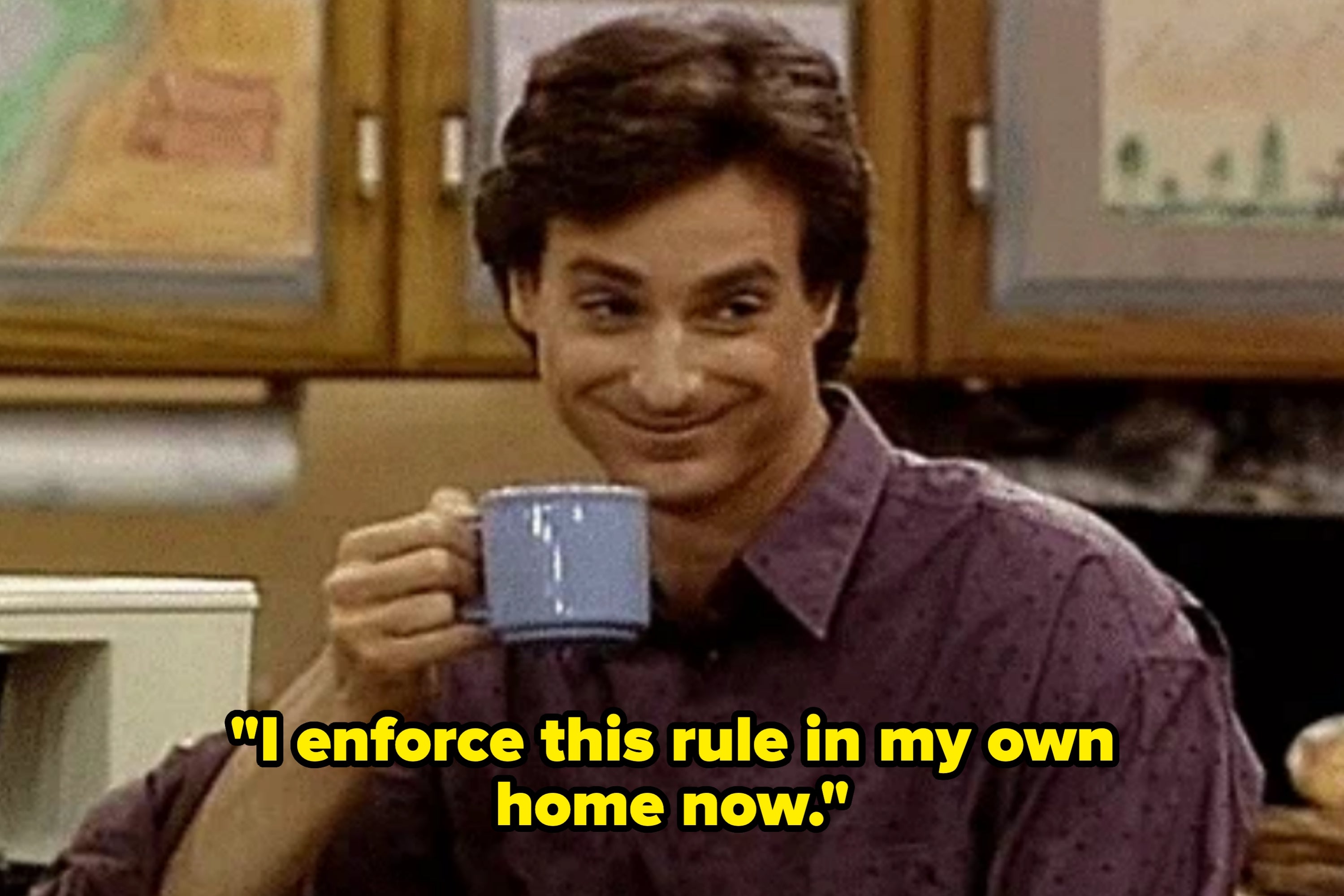Person in a patterned shirt smirking while holding a mug, seated in a kitchen setting