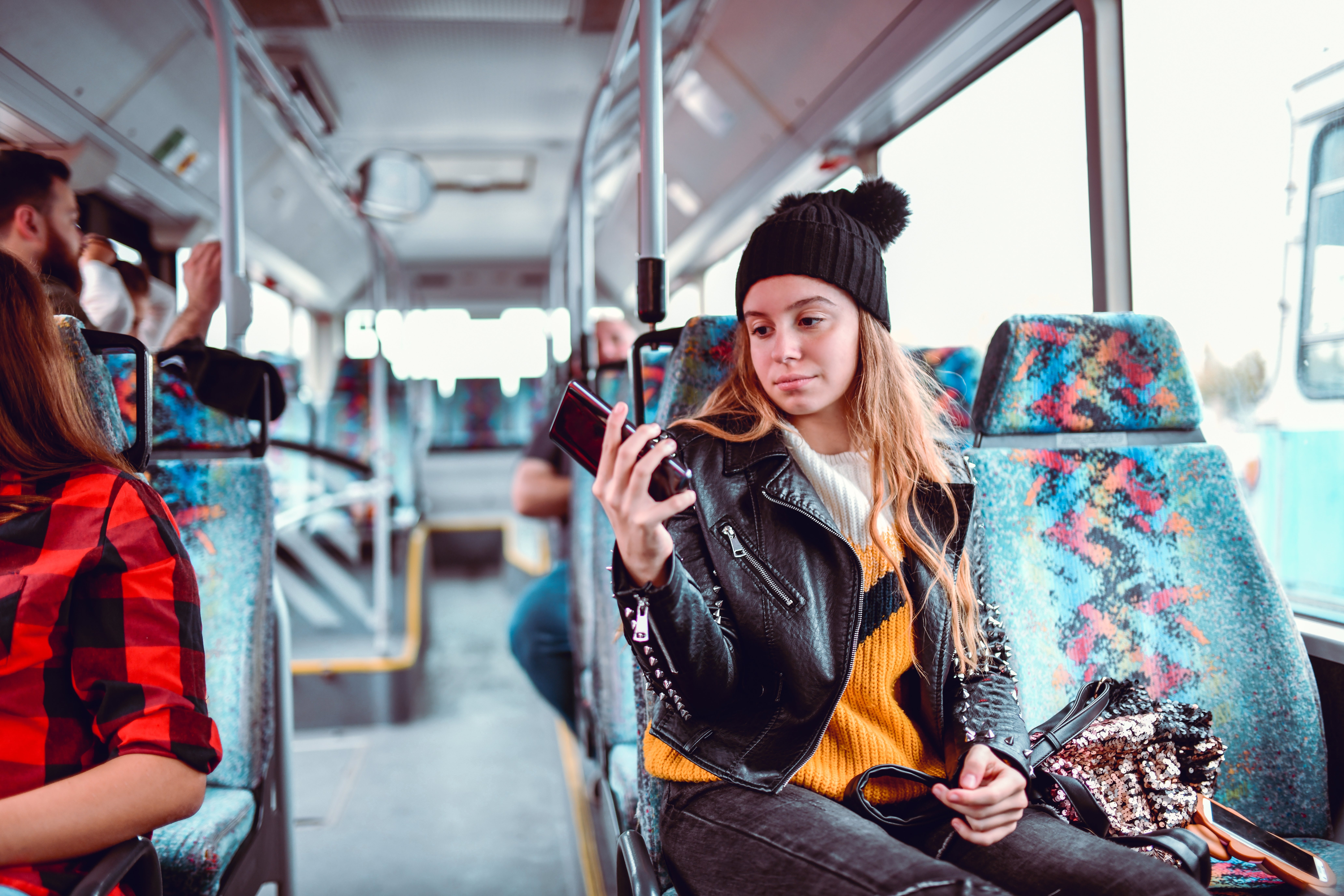Teenager successful  a beanie and leather overgarment   looks astatine  her telephone  connected  a bus, surrounded by passengers