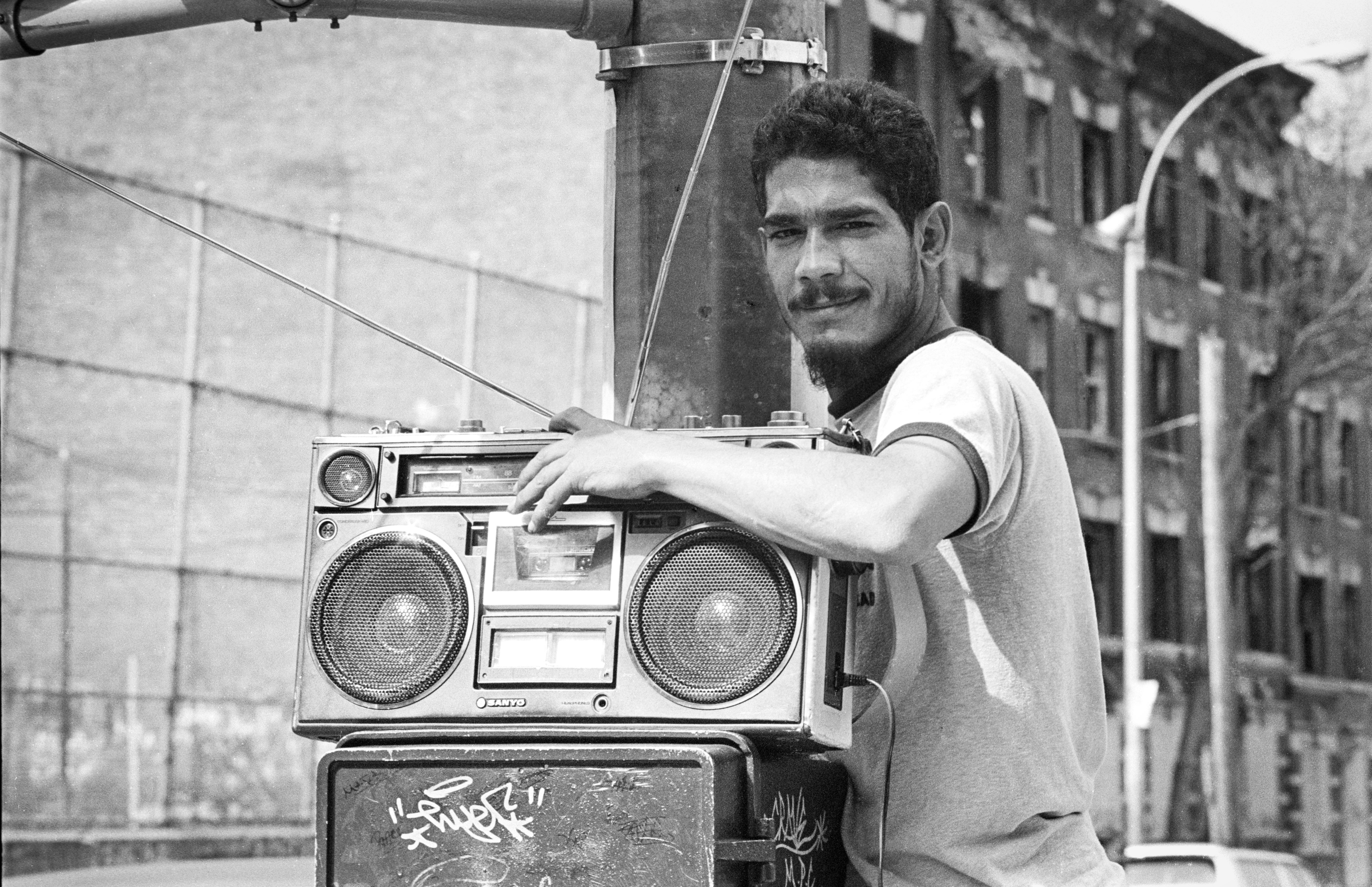 A antheral   successful  casual attire leans connected  a ample  boombox connected  a thoroughfare  corner, expressing a relaxed and assured  demeanor
