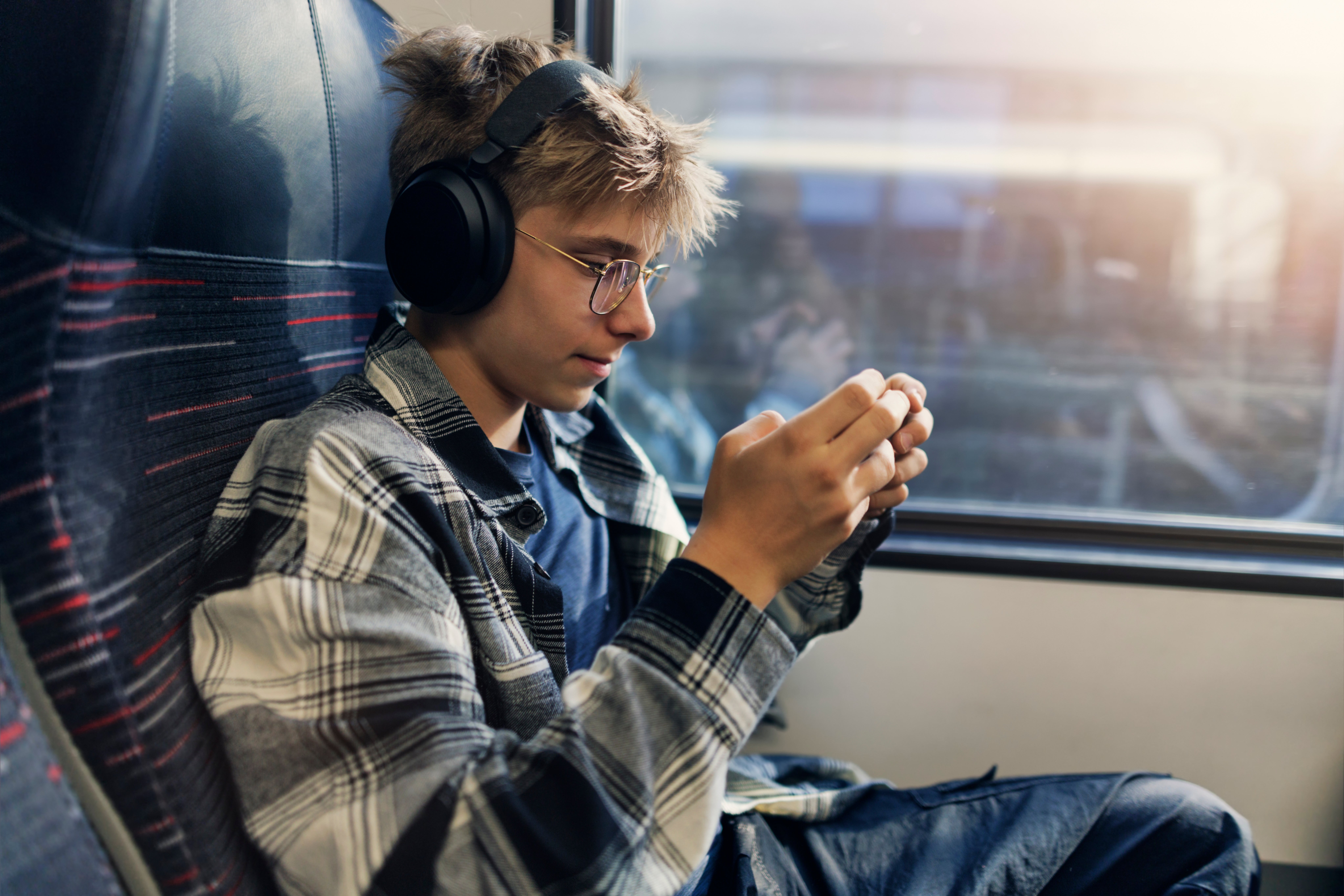 Teen wearing headphones and glasses, focused connected  a smartphone, sits connected  a bid     seat. Casual attire with a plaid shirt