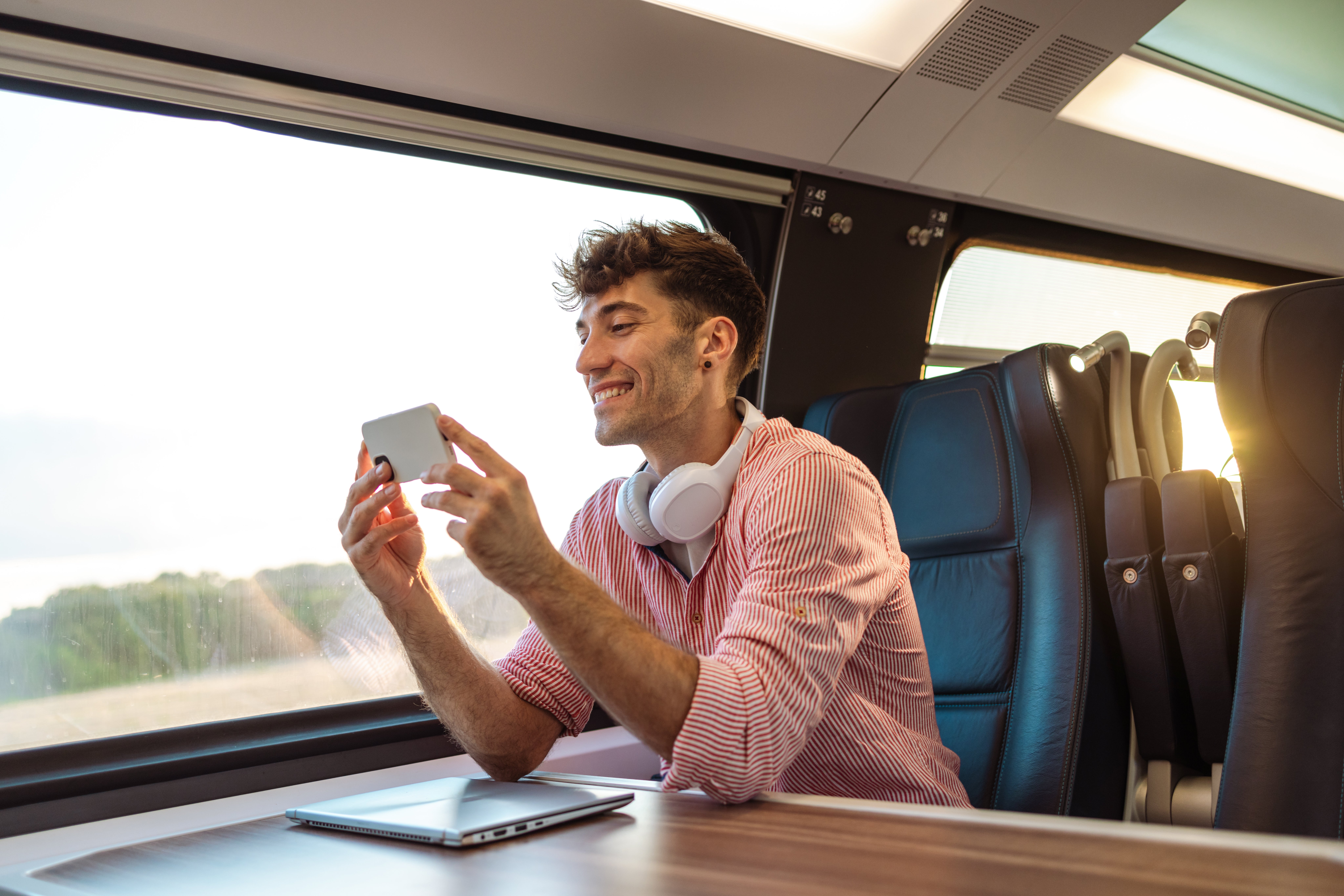 Smiling antheral   connected  a train, wearing headphones, looking astatine  his phone. A laptop is connected  the array  successful  beforehand   of him