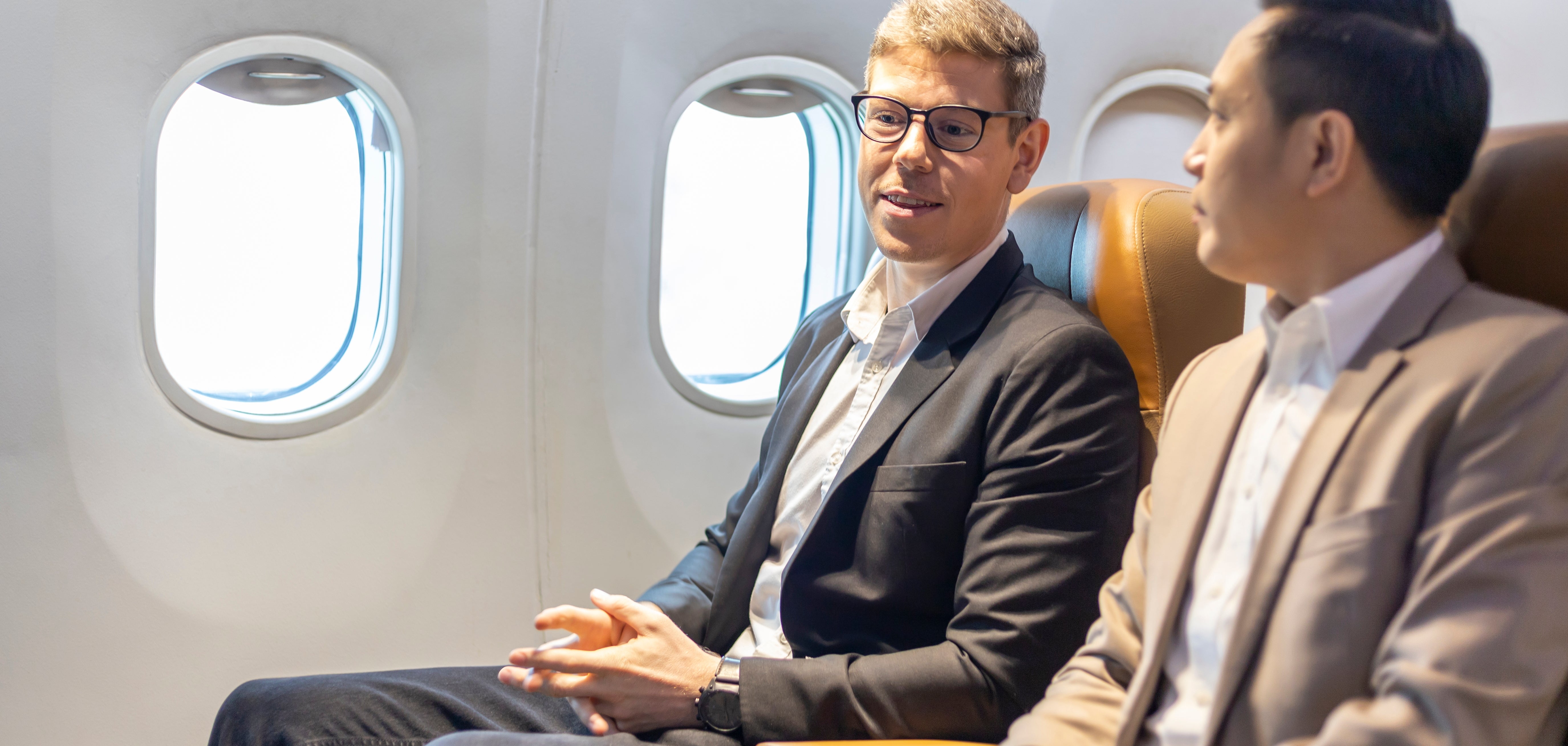 Two men successful  concern  attire speech   portion    seated successful  an airplane, looking engaged and relaxed