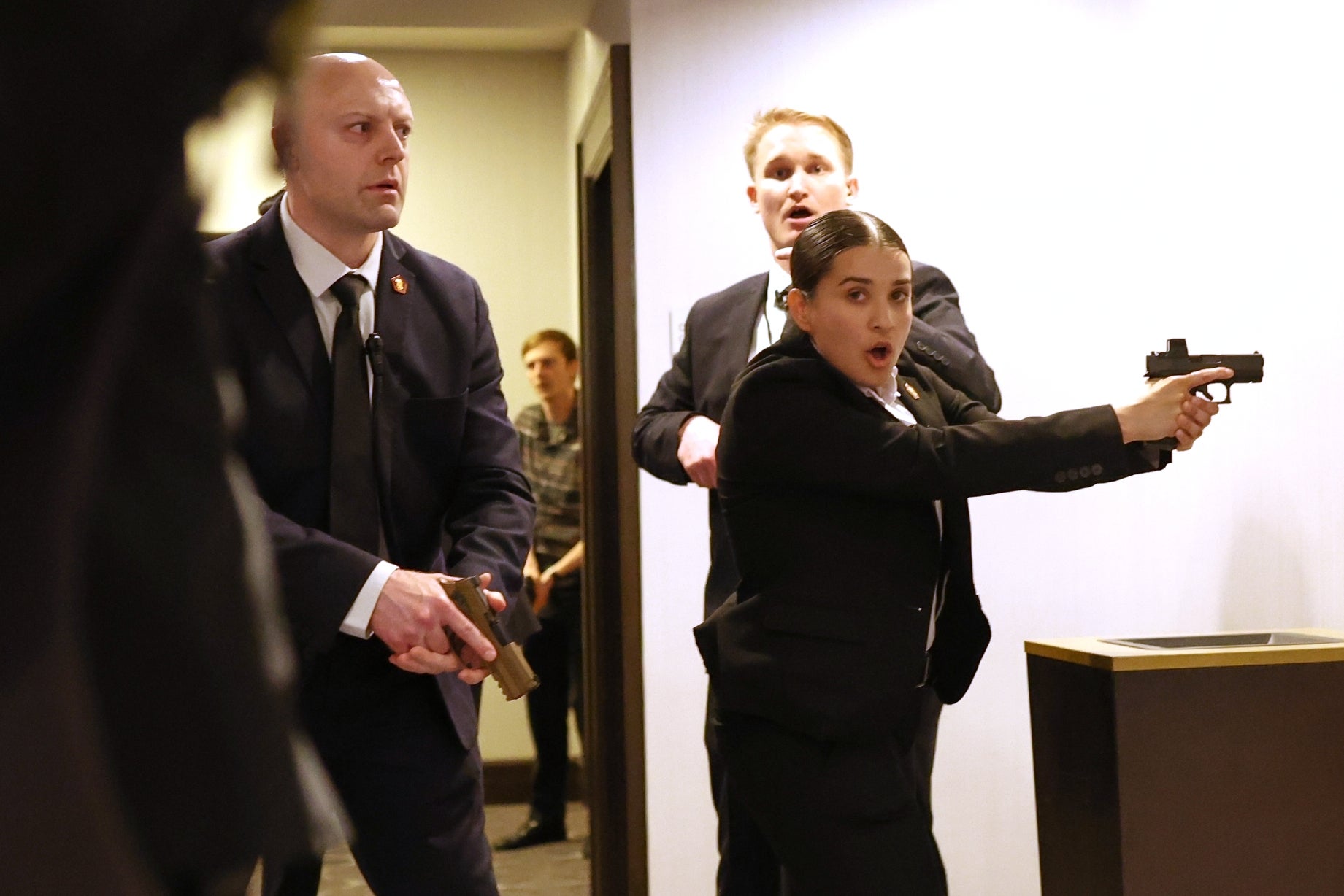 Individuals in suits in a tense scene, one pointing a handgun, appearing to be in a security or law enforcement role