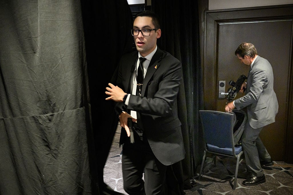 A formally dressed individual in a suit walks quickly through a curtain, followed by another person carrying equipment