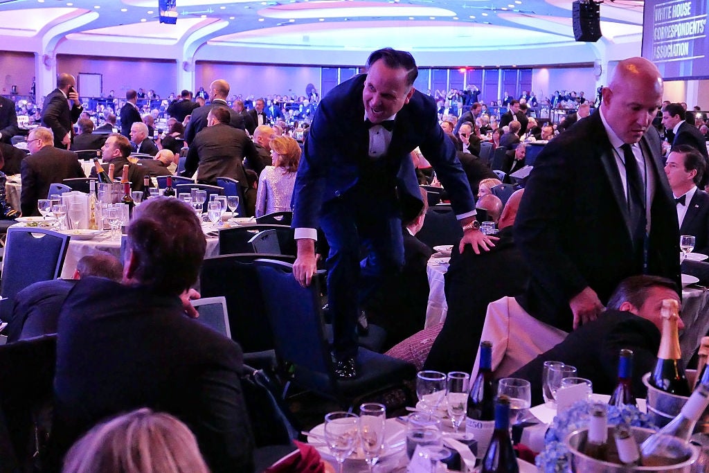 A man in formal wear climbs over banquet chairs amid a crowded event setting, capturing attention with his unusual movement