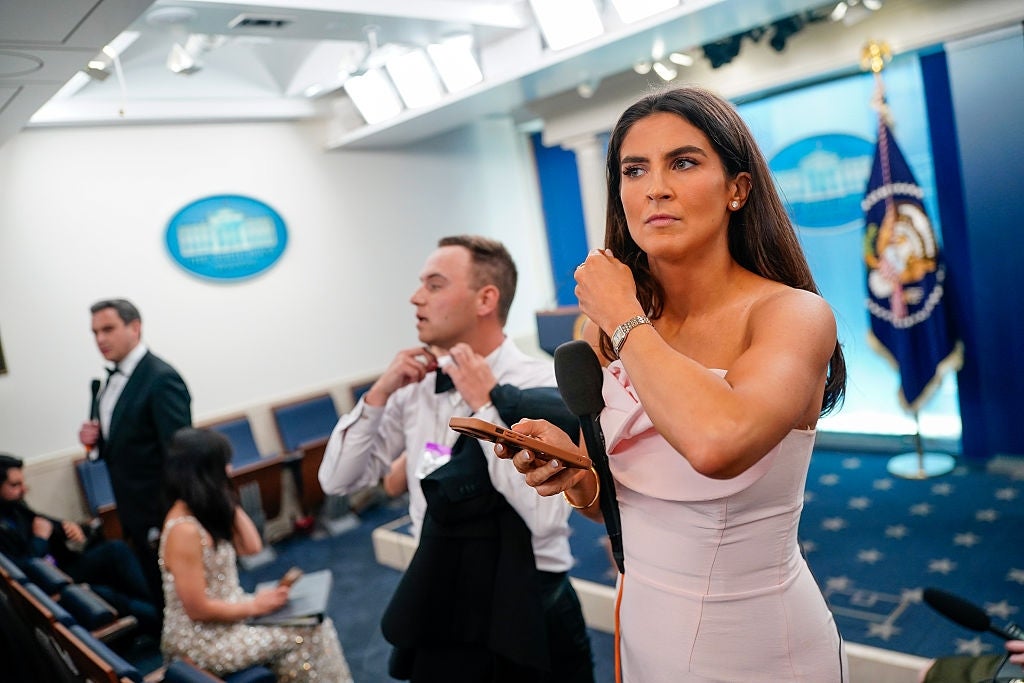 Person in formal attire prepares for a press event at a government venue, holding a microphone and phone. Staff adjust clothing in the background