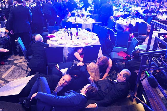 People in formal attire lie on the ground under tables at a formal event, appearing to take cover or participate in a playful activity