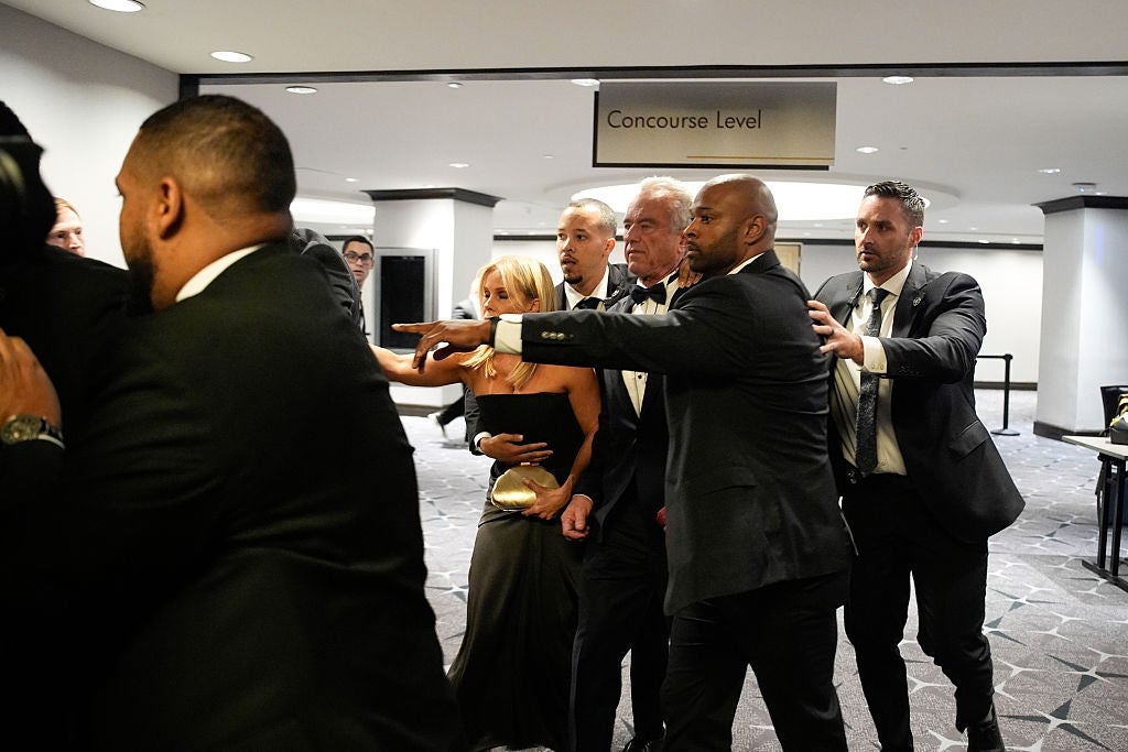 A group of formally dressed people are in a lobby, some pointing and appearing to guide others. One person is holding a woman's arm