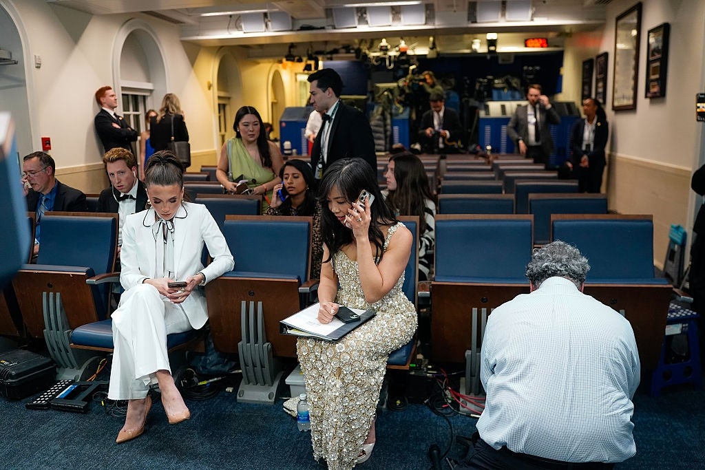Journalists in a briefing room, some taking notes and others on laptops, await a press conference's start. Two seated women focus on phones