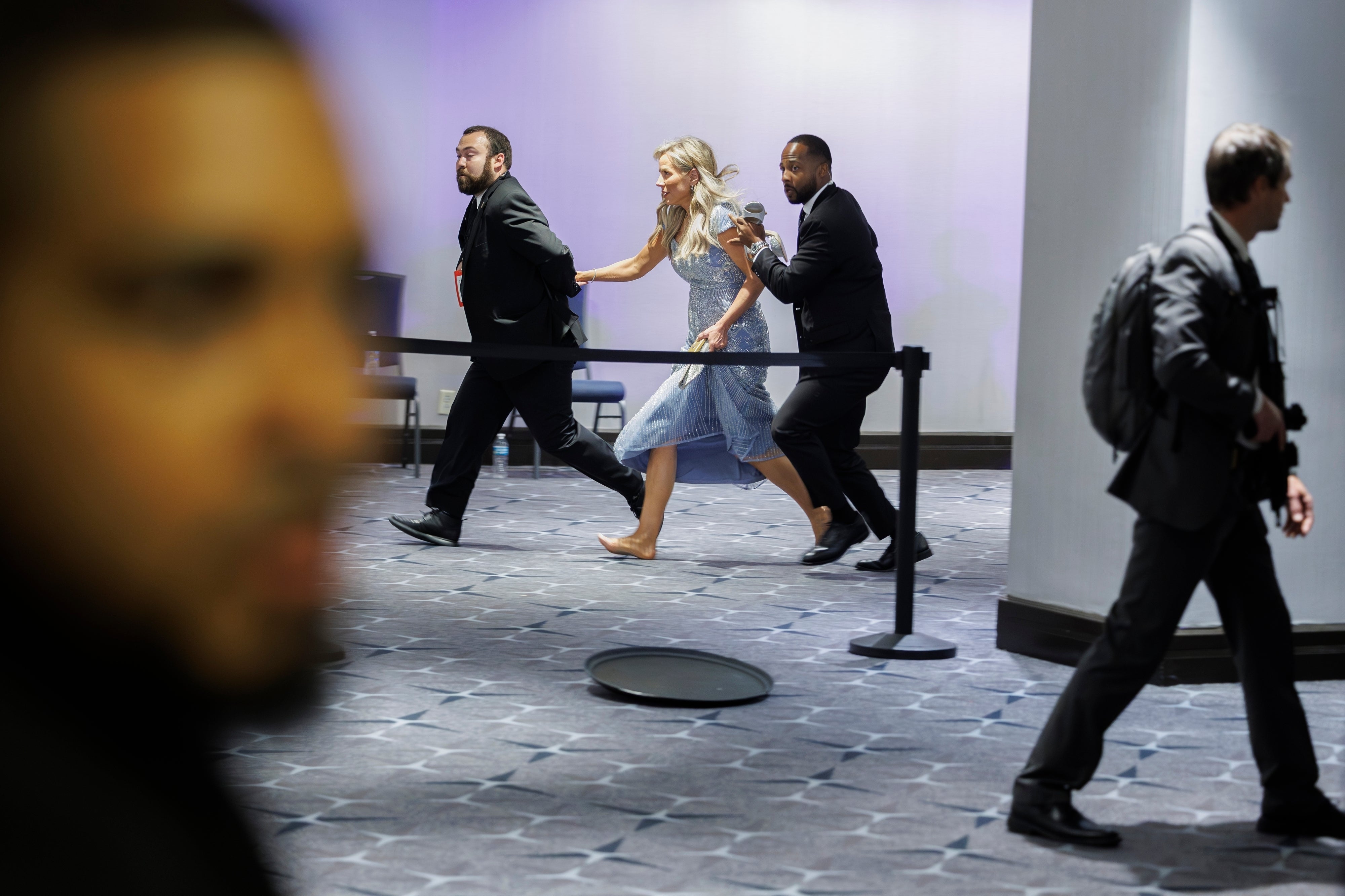Person in elegant dress and heels running behind security personnel in a public space; another person walks nearby, blurred foreground