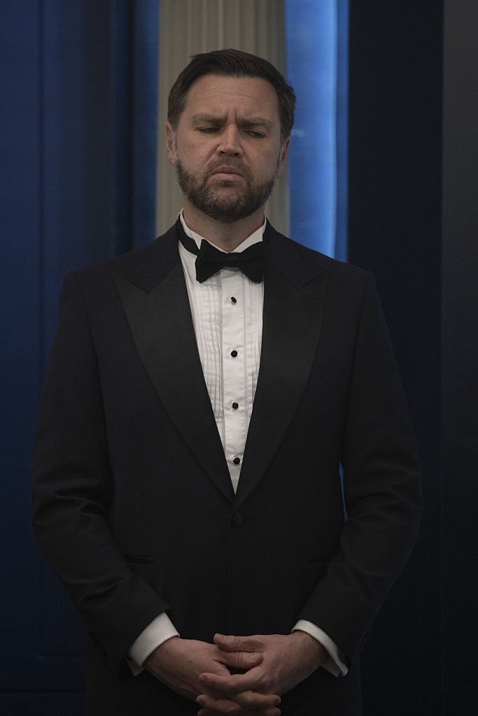 A man in a tuxedo with a bowed head and clasped hands stands indoors, appearing contemplative