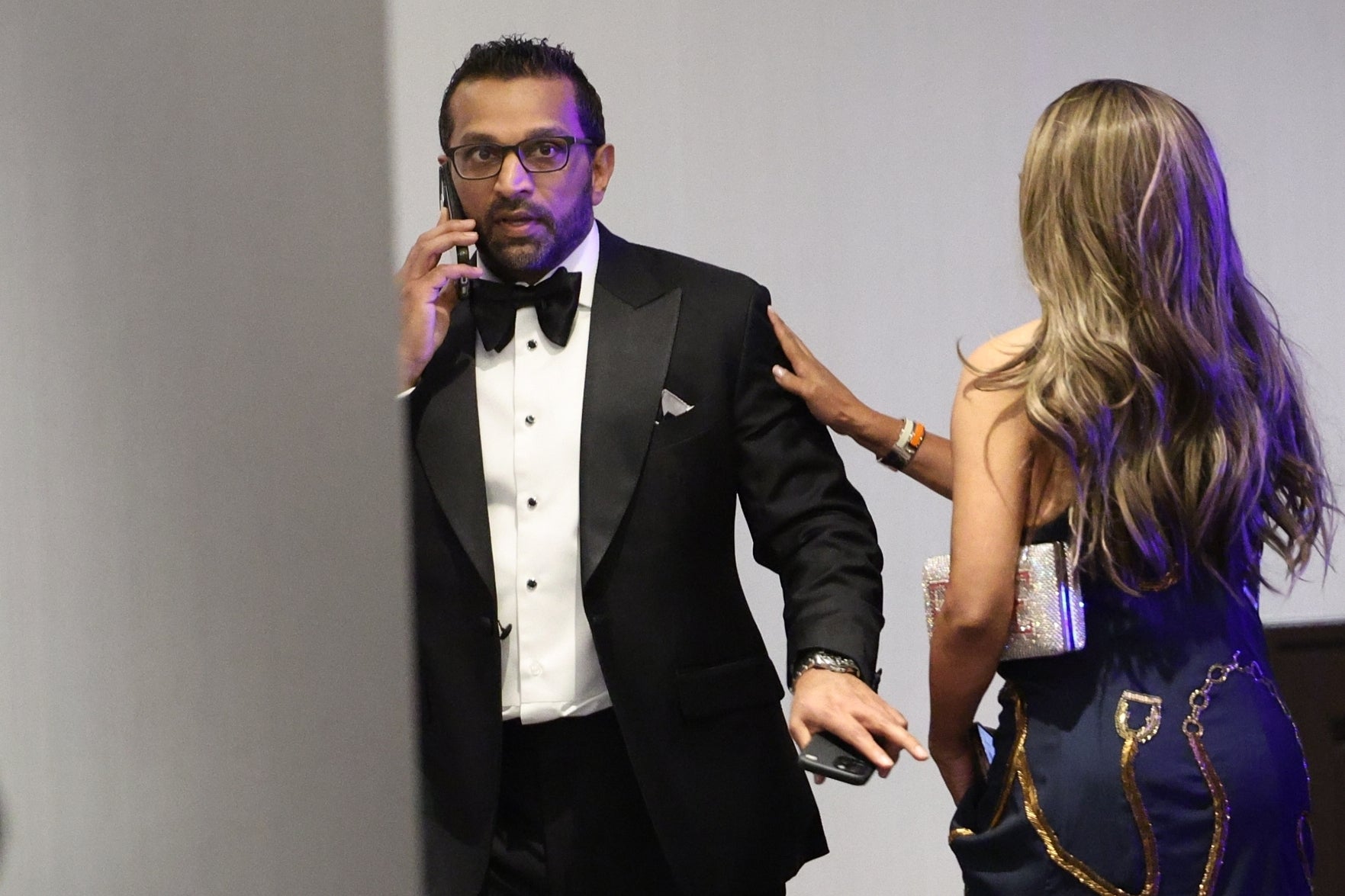 Man in tuxedo on phone, looking surprised, accompanied by a woman in elegant dress holding his arm