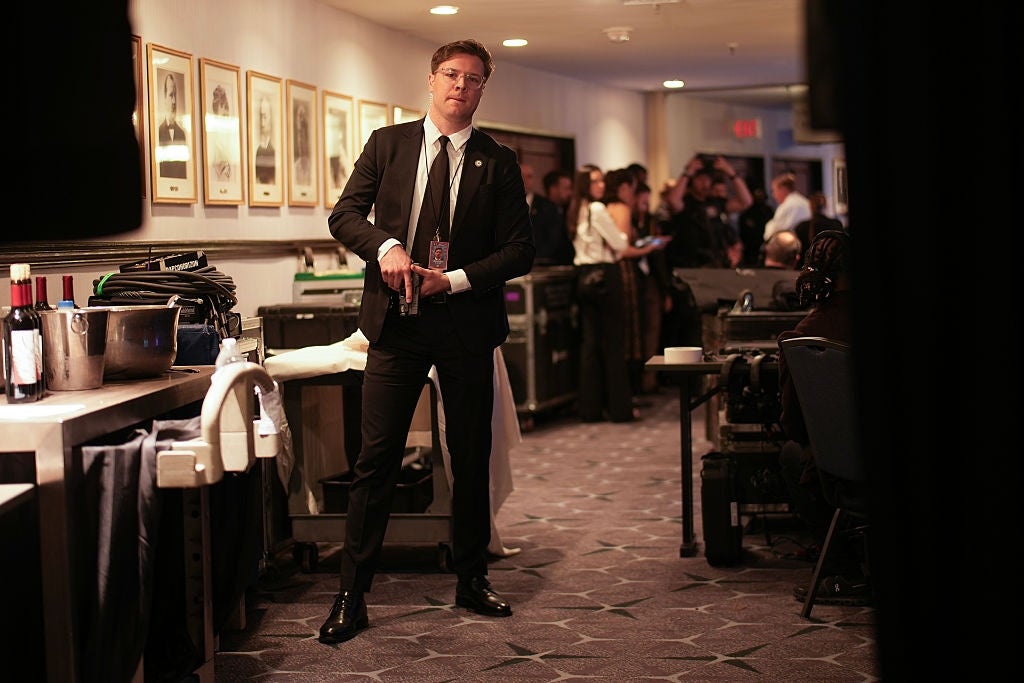 A person in a suit holds a bottle backstage at an event, with framed photos and people in the background