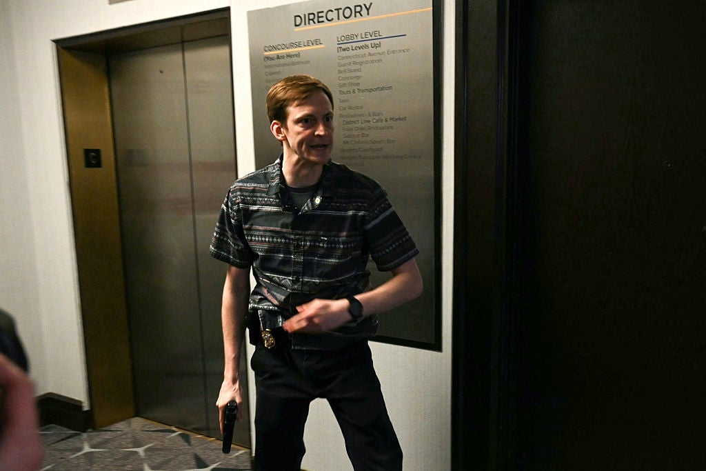 A man wearing a striped shirt and dark pants stands in a hallway, holding a tablet and gesturing in a conversation