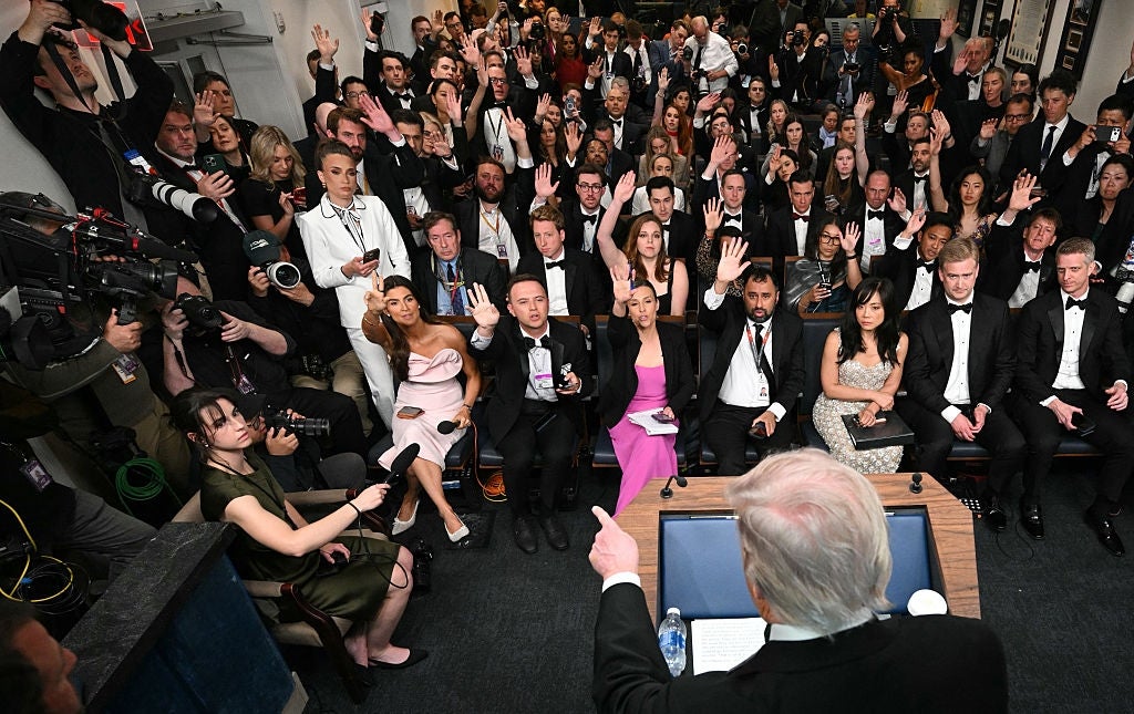 Crowded press room with a speaker at the podium and audience members raising hands, dressed formally, possibly at a press event or awards ceremony