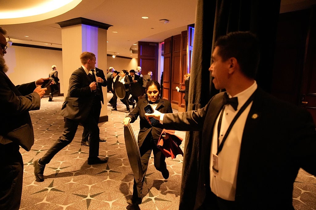 People in formal attire run in a hotel lobby during an event, possibly a drill or emergency. A security person holds back a curtain