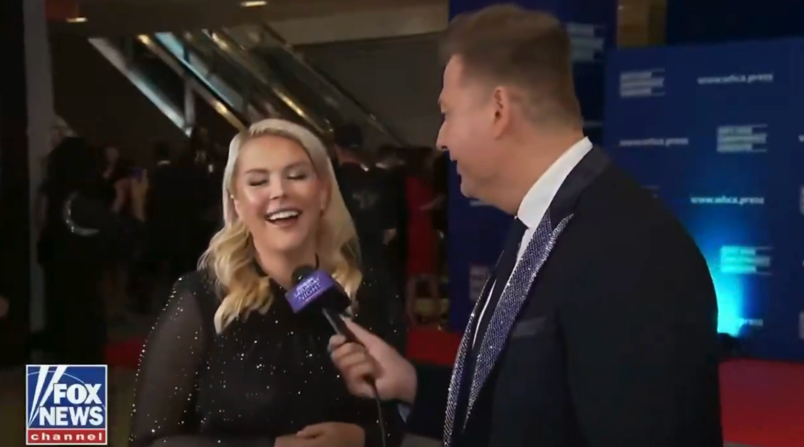 A pistillate   successful  a sheer, glittery formal  is being interviewed connected  a reddish  carpet by a antheral   successful  a suit. The backdrop has logos and a reddish  carpet setting