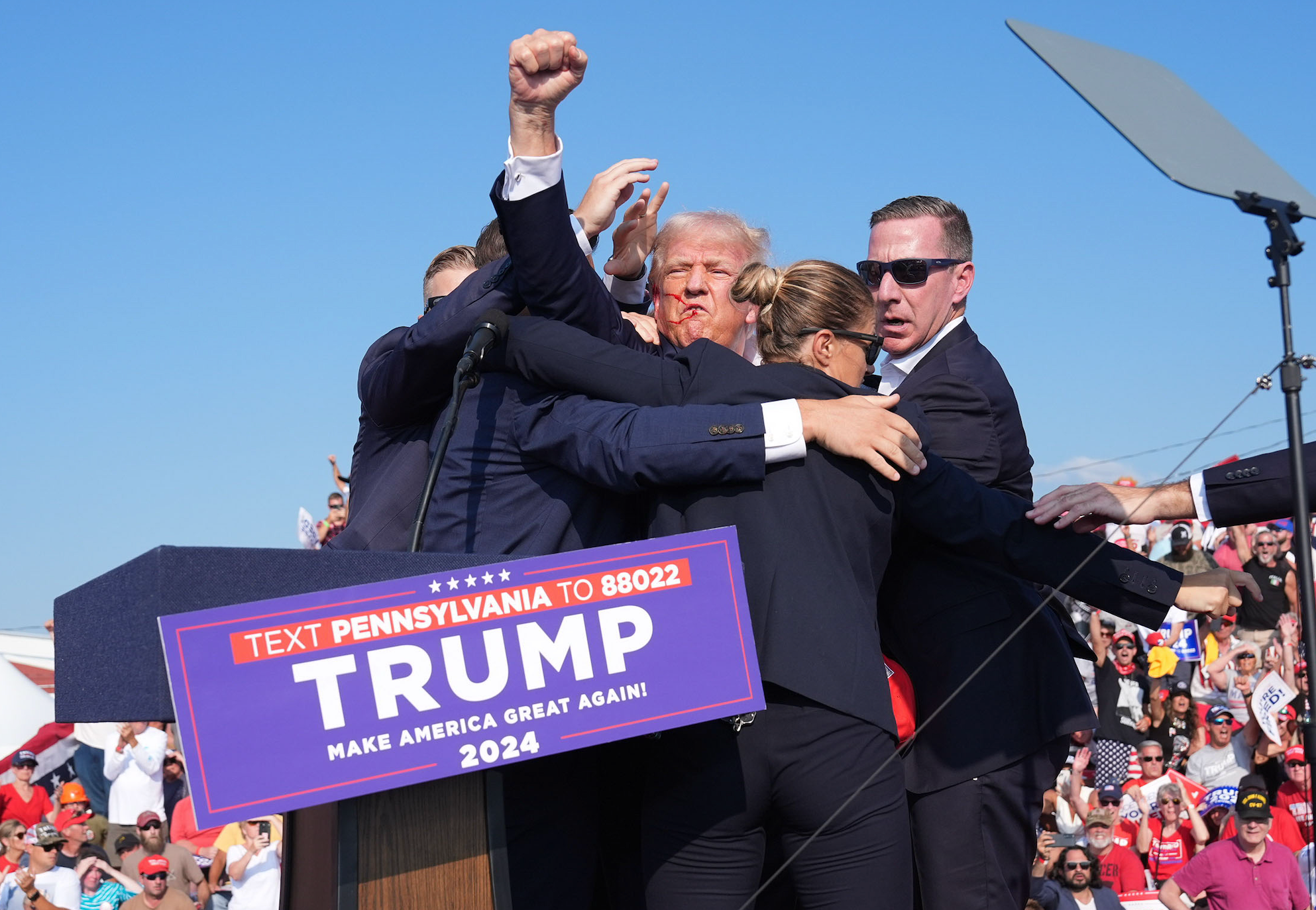 En gruppe sikkerhetspersonell omringer Donald Trump på scenen ved et offentlig arrangement. Et skilt lyder: 