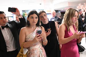 Guests in formal attire, holding smartphones, appear focused at an event, suggesting a news or media gathering
