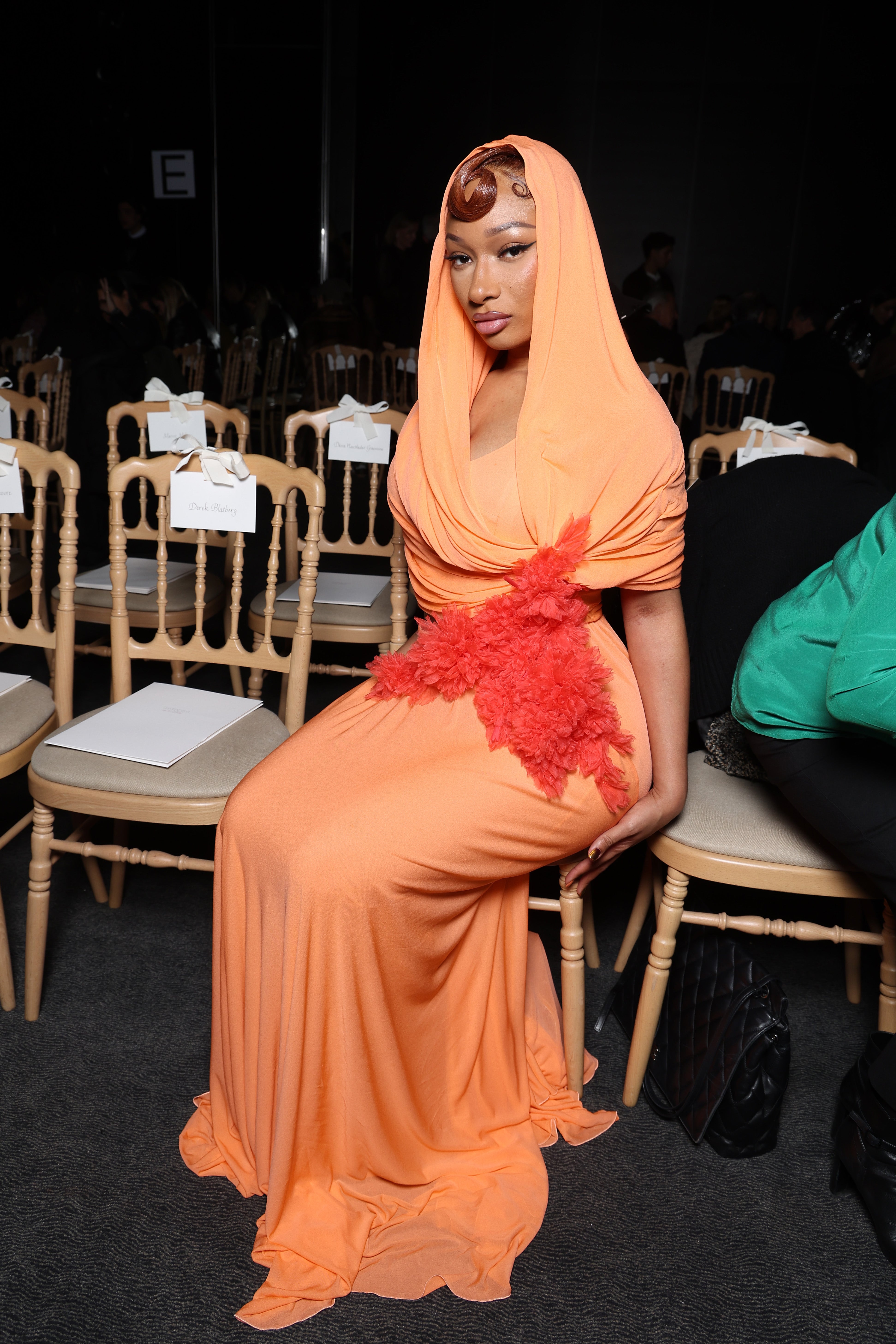Megan Thee Stallion posing successful  an elegant floor-length gown with a draped hood and reddish  floral detail, sitting connected  a seat  astatine  a ceremonial  event