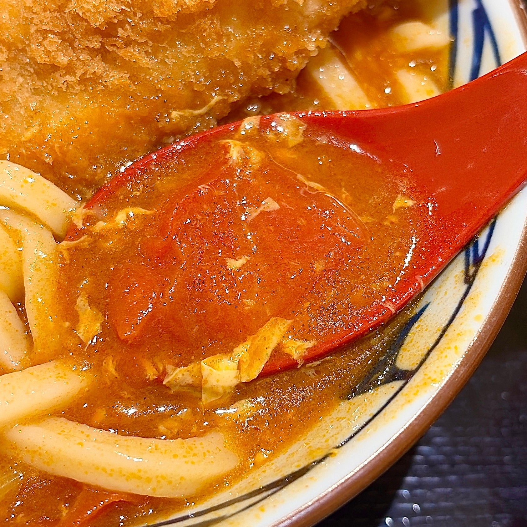 丸亀製麺「海老カツトマたまカレーうどん」うどんの麺とカレーソースが入ったどんぶりに、赤いスプーンが盛られている。