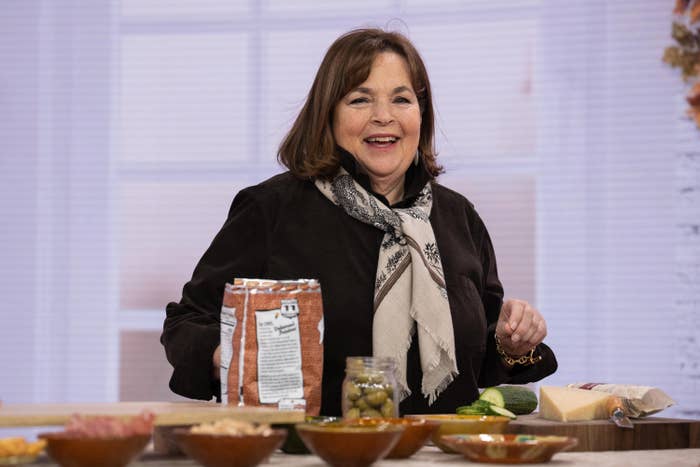 A pistillate   smiles portion    cooking, holding a container  of flour beside ingredients connected  a counter, wearing a scarf and a acheronian  outfit