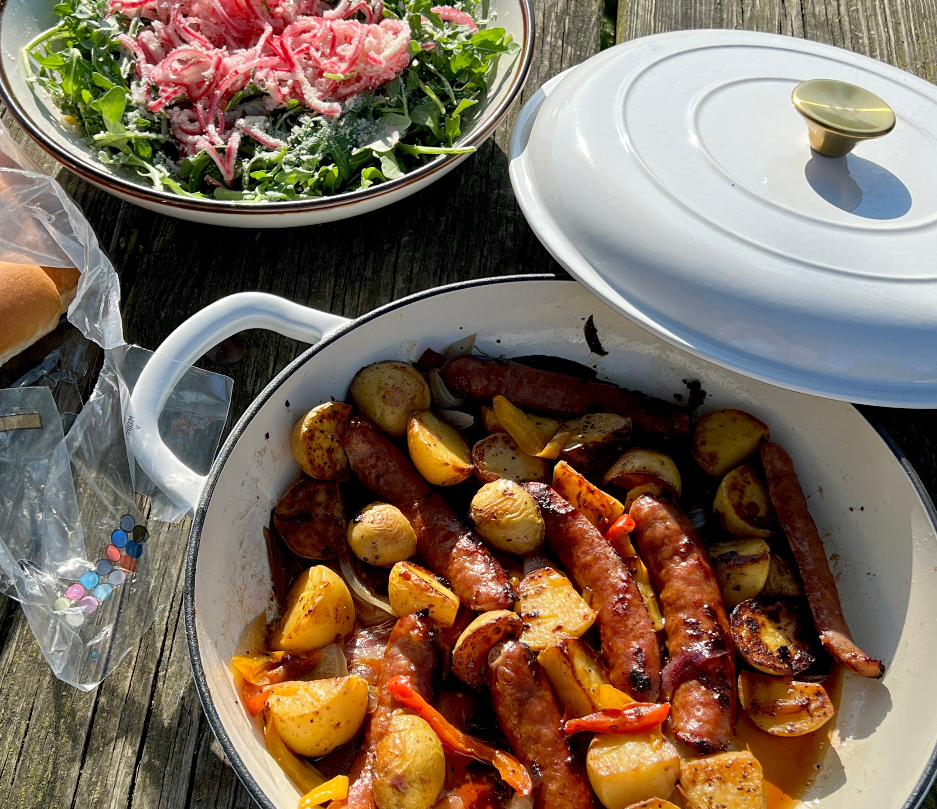 A pan of grilled sausages and potatoes next to a bowl of leafy salad with a pink topping on a wooden picnic table