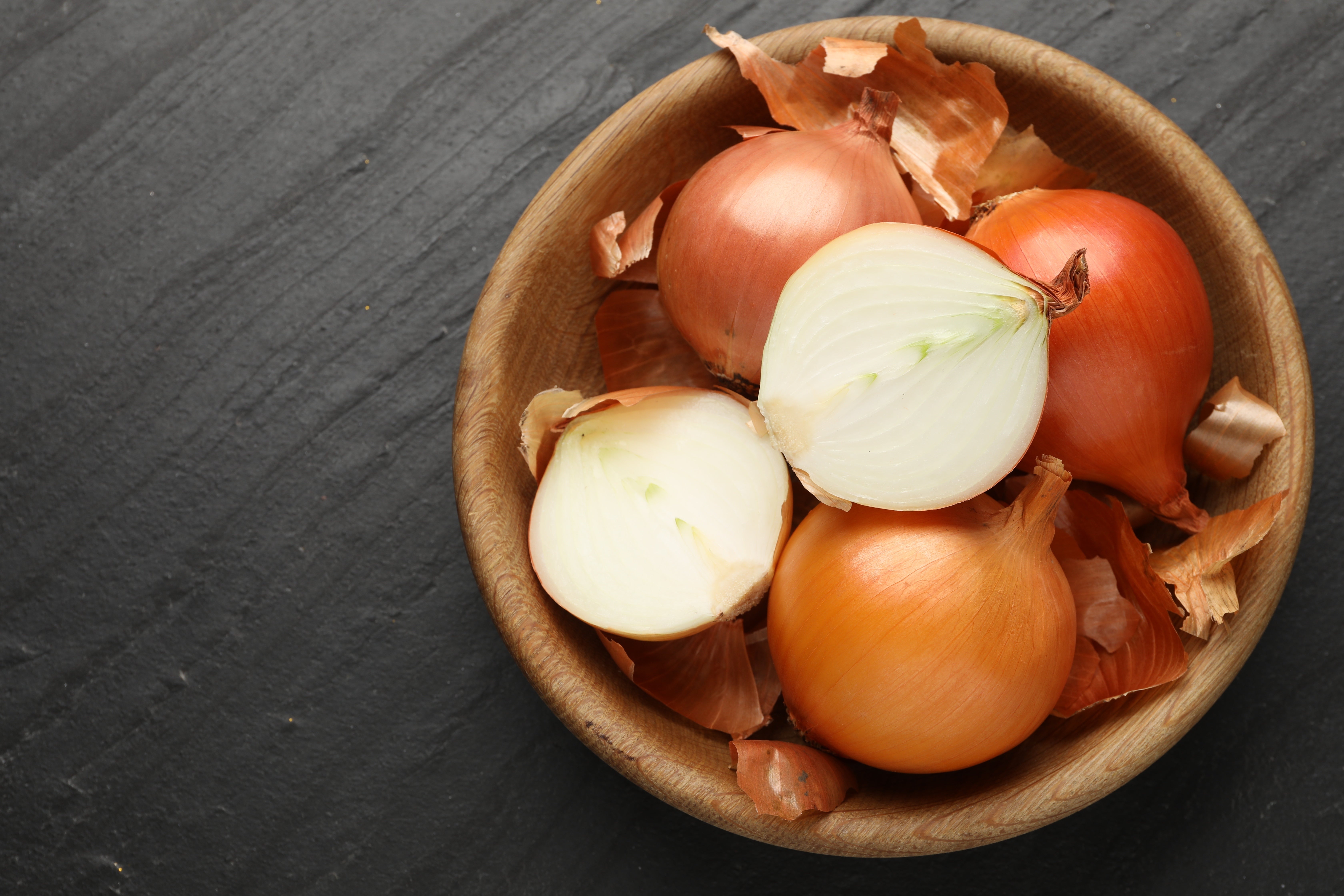 A wooden bowl filled with whole and halved onions on a dark slate surface