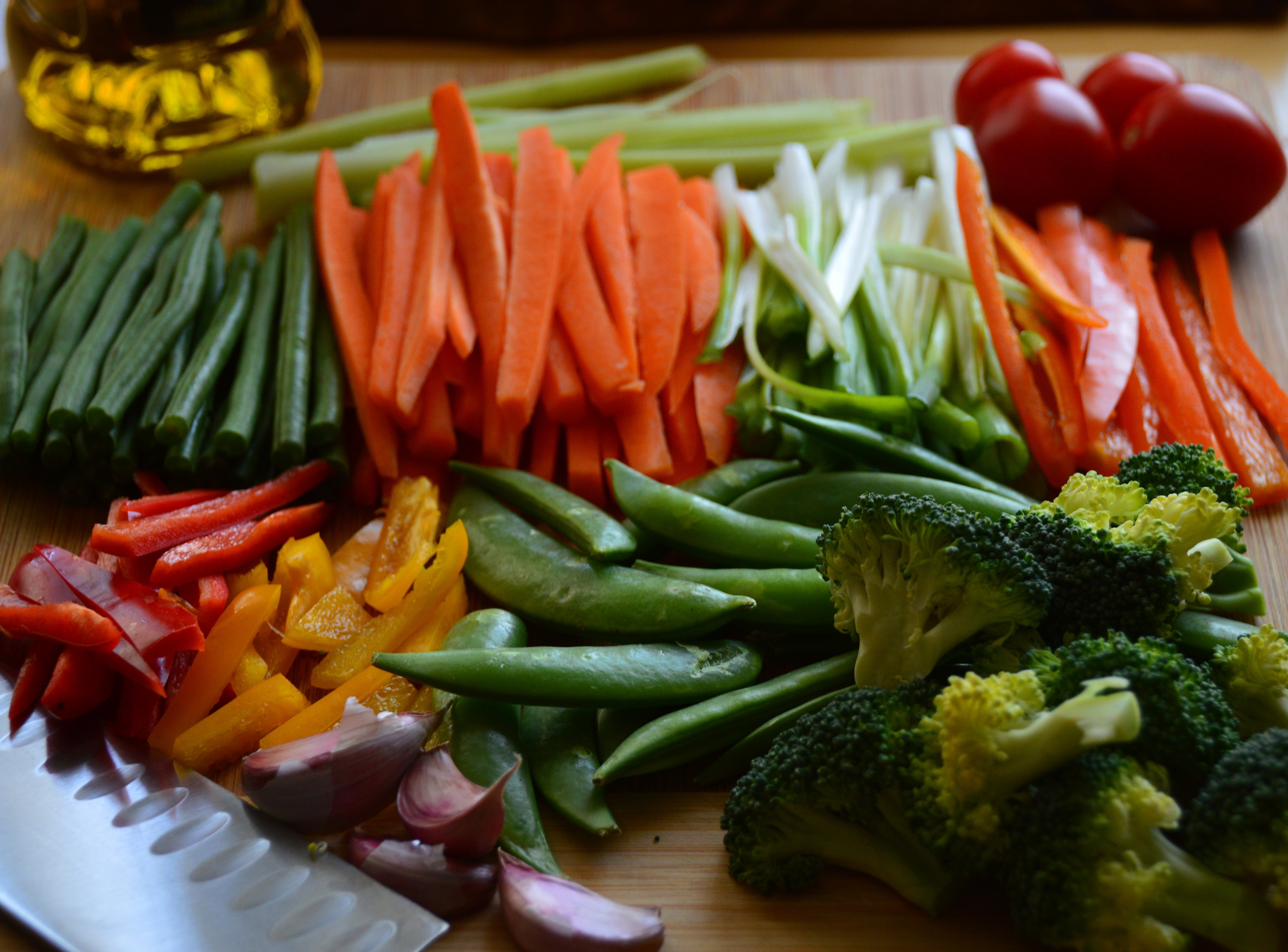 An assortment of fresh vegetables, including carrots, bell peppers, broccoli, green beans, and tomatoes, are sliced and placed on a wooden board