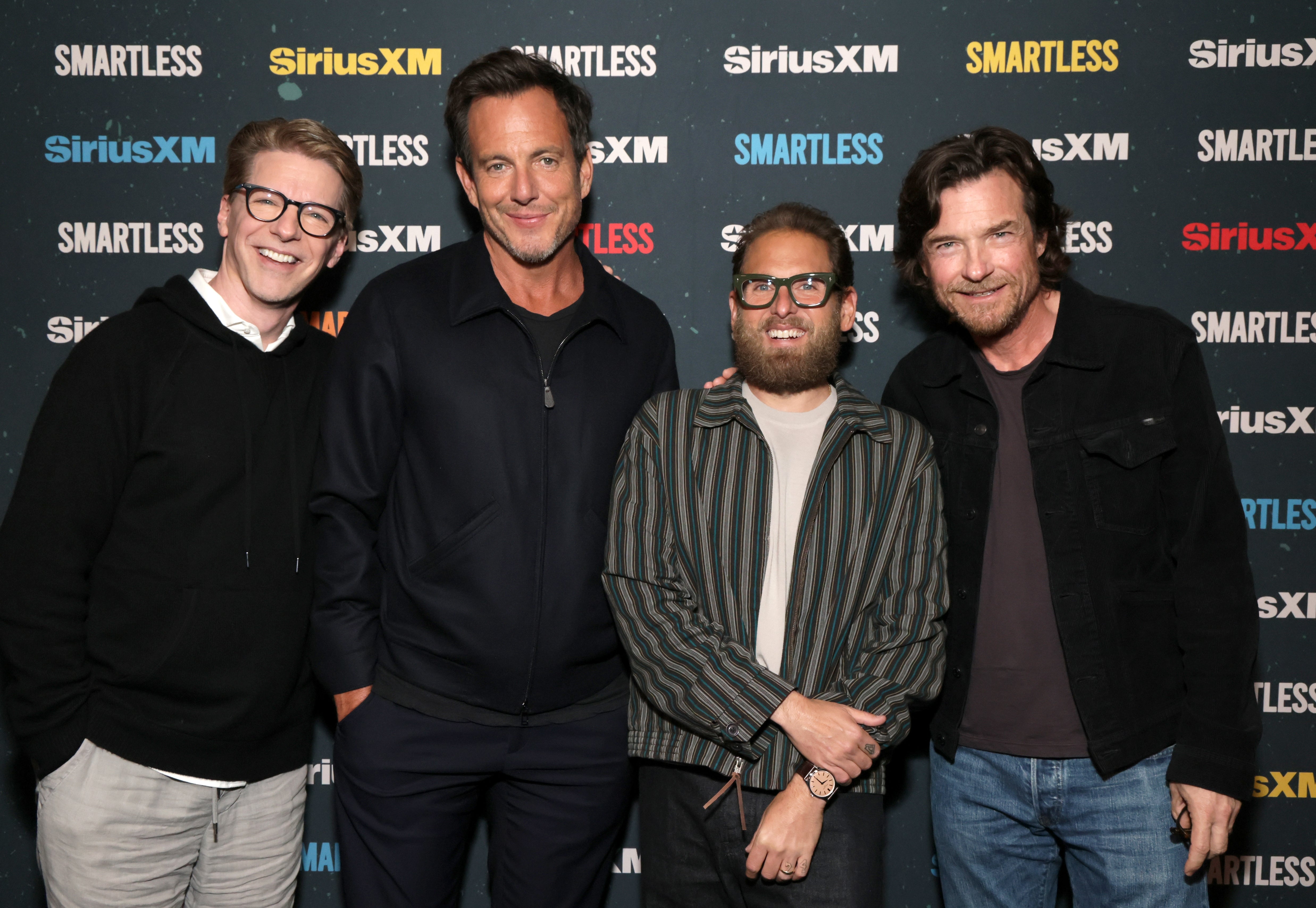 Four radical   basal   unneurotic  astatine  a SiriusXM and SmartLess event. They are casually dressed, immoderate   wearing glasses and jackets, smiling for the photo