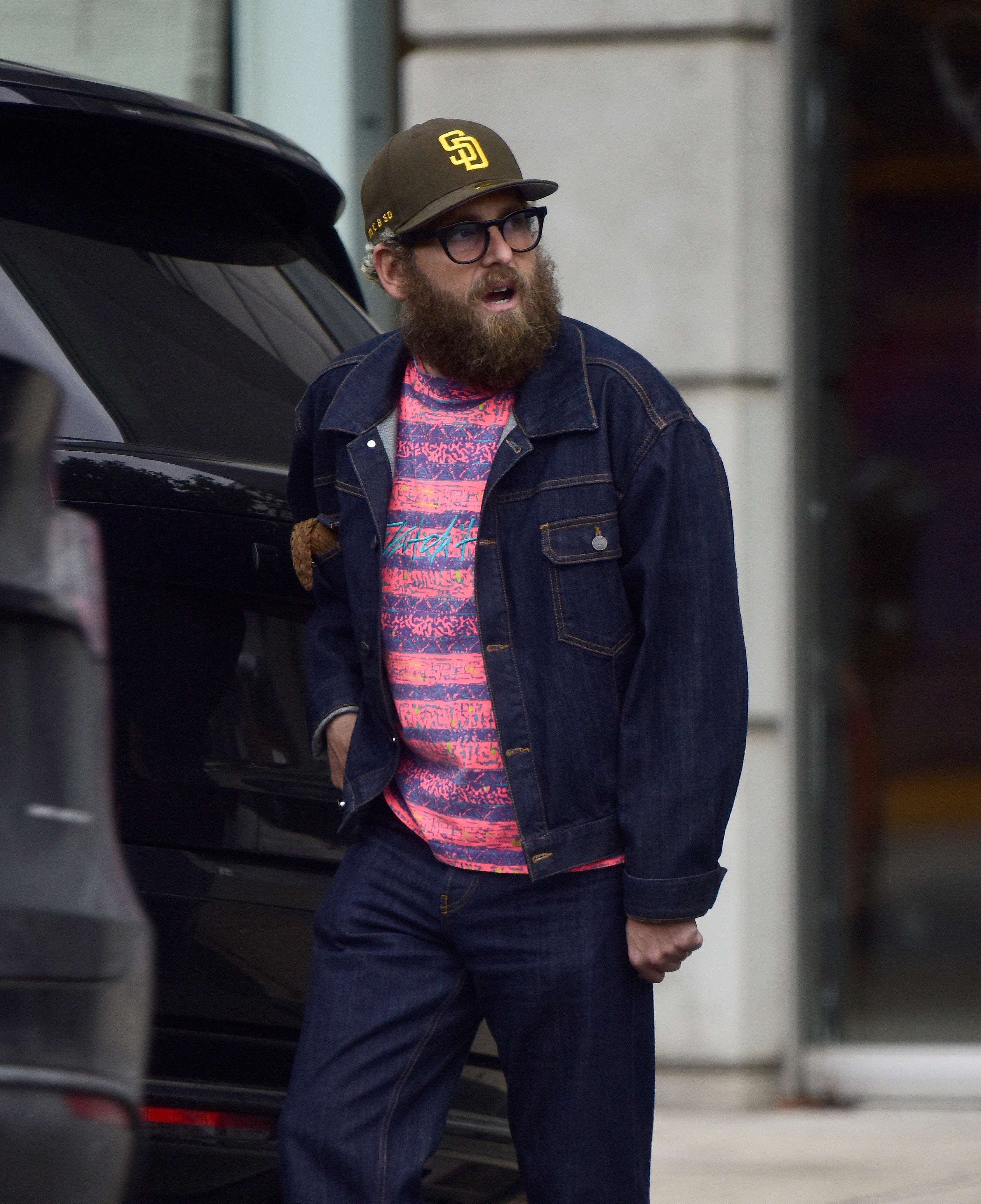 Person wearing a denim jacket, agleam  patterned shirt, jeans, and a shot   cap, lasting  adjacent   a vehicle