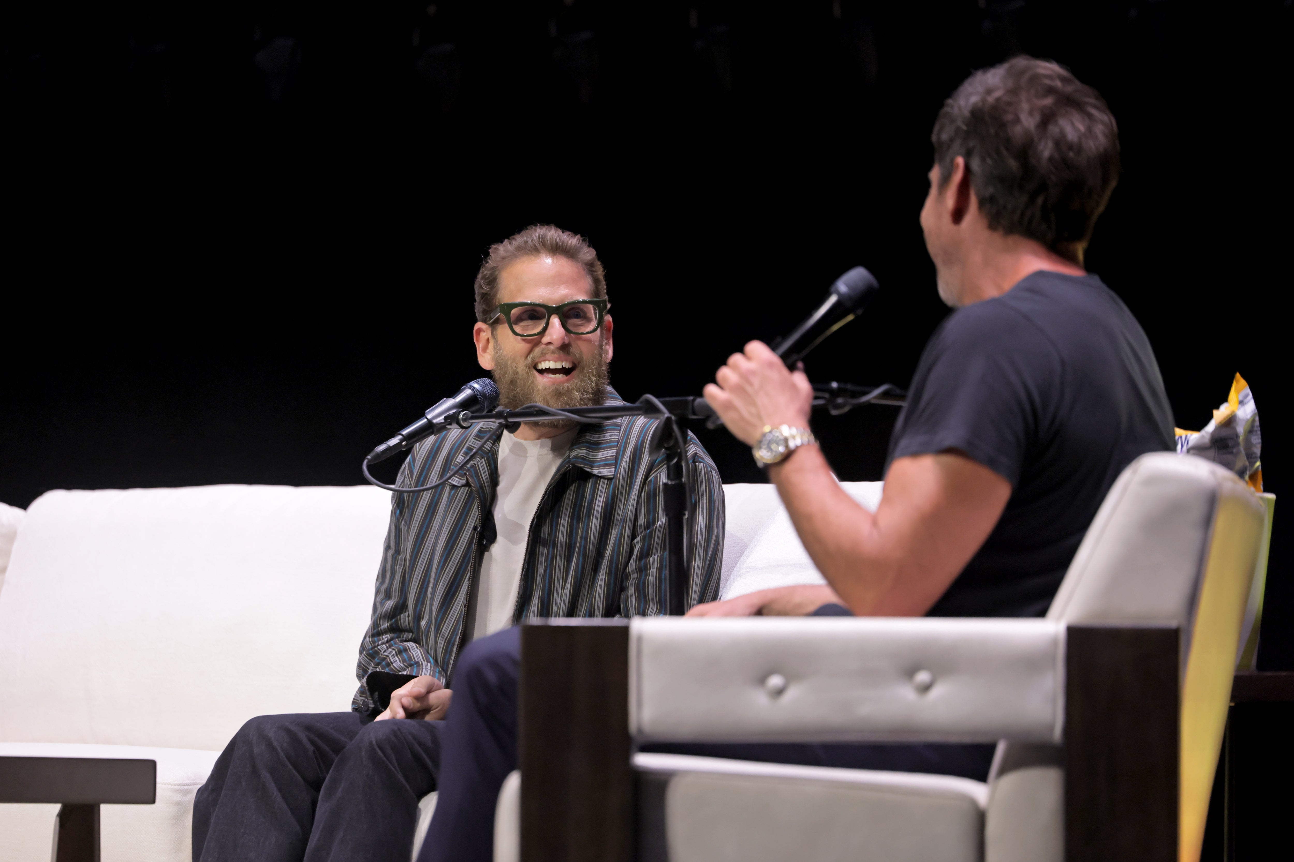 Two radical   beryllium   connected  signifier    having a conversation. One wears a striped overgarment   and glasses. Both clasp  microphones, smiling arsenic  they talk