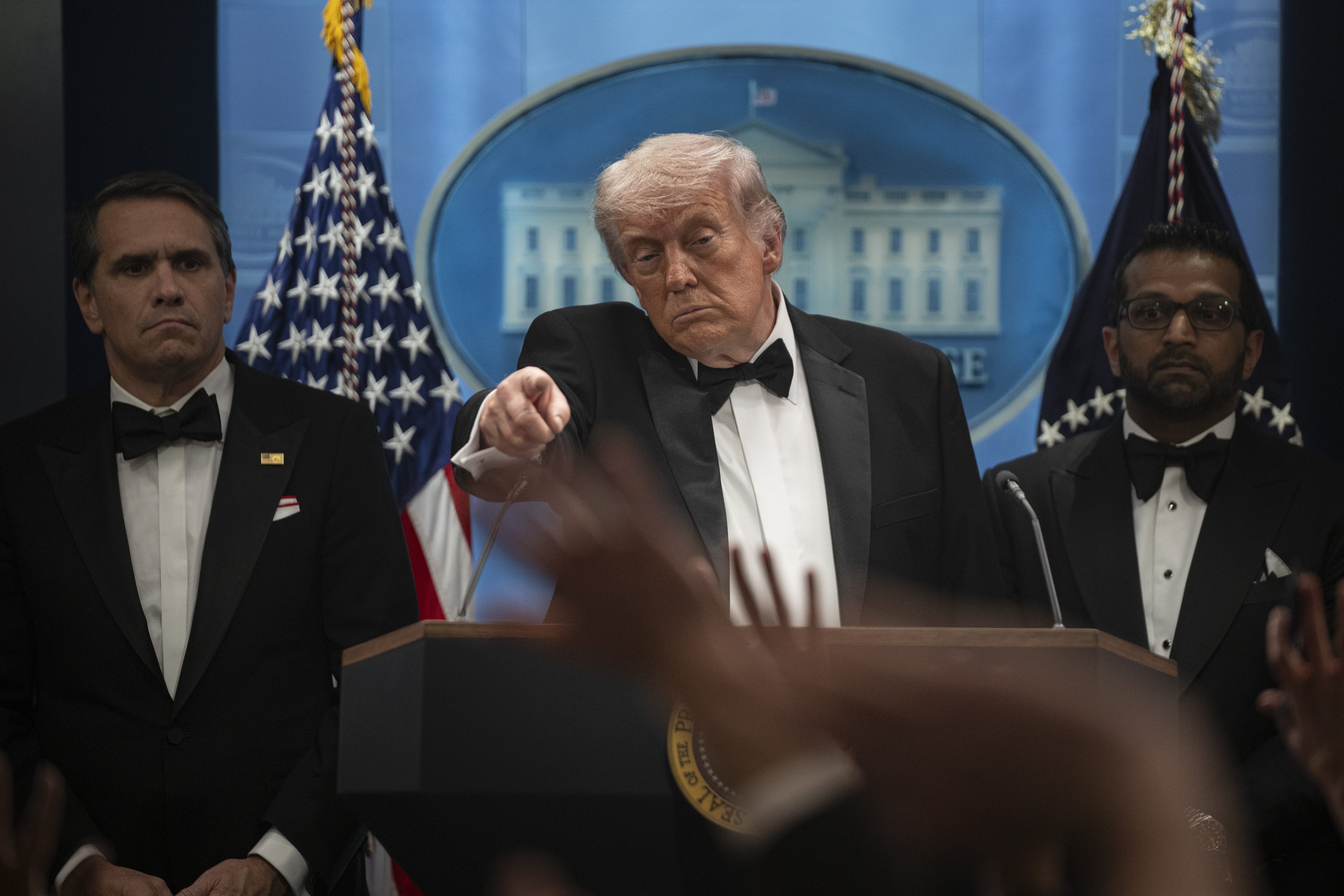 A idiosyncratic   successful  a tuxedo gestures portion    speaking astatine  a podium, flanked by 2  individuals successful  tuxedos, with a backdrop featuring the White House emblem