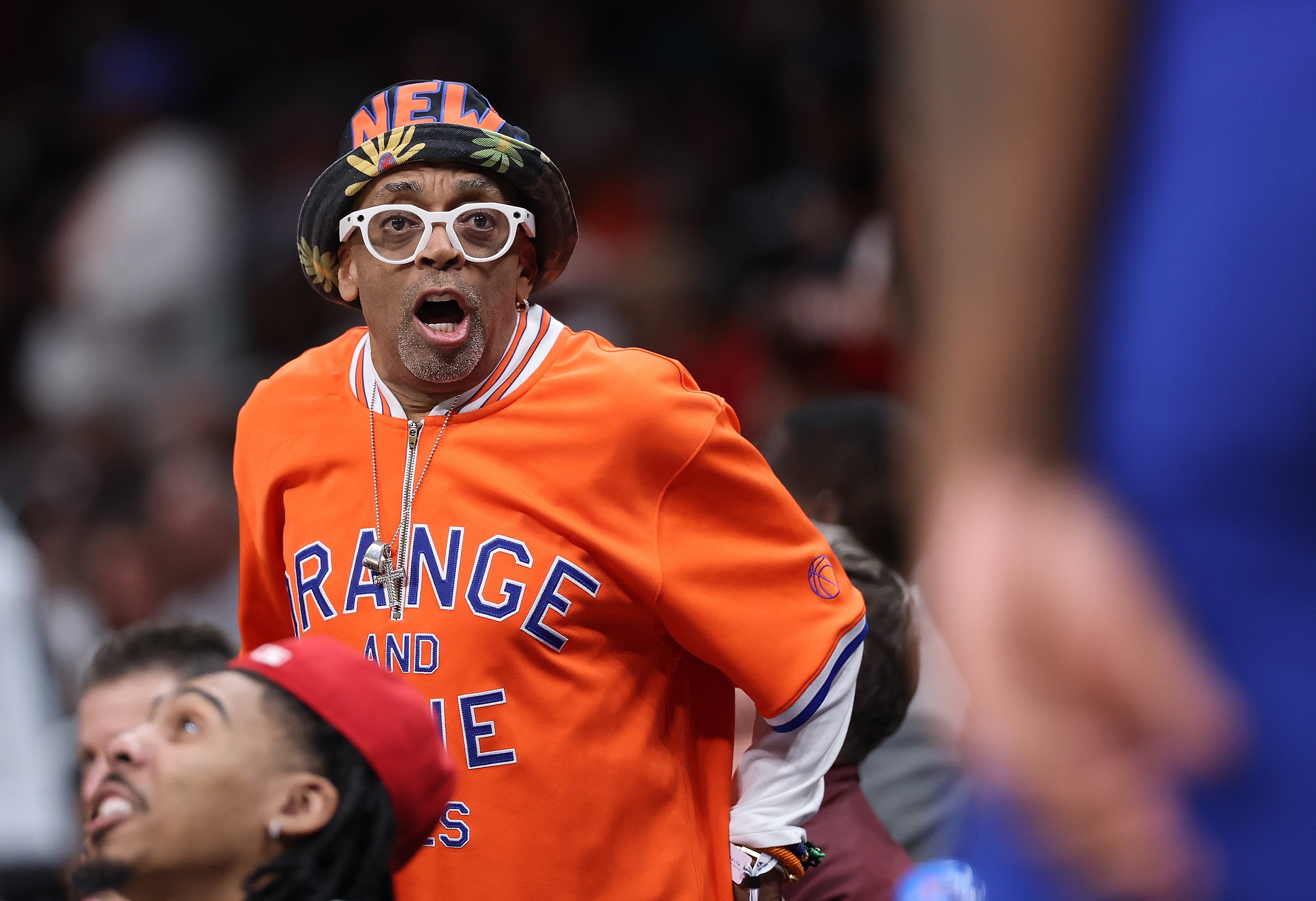 A idiosyncratic   enthusiastically reacts astatine  a sports event, wearing a hat, glasses, and a casual orangish  garment  with substance   connected  it