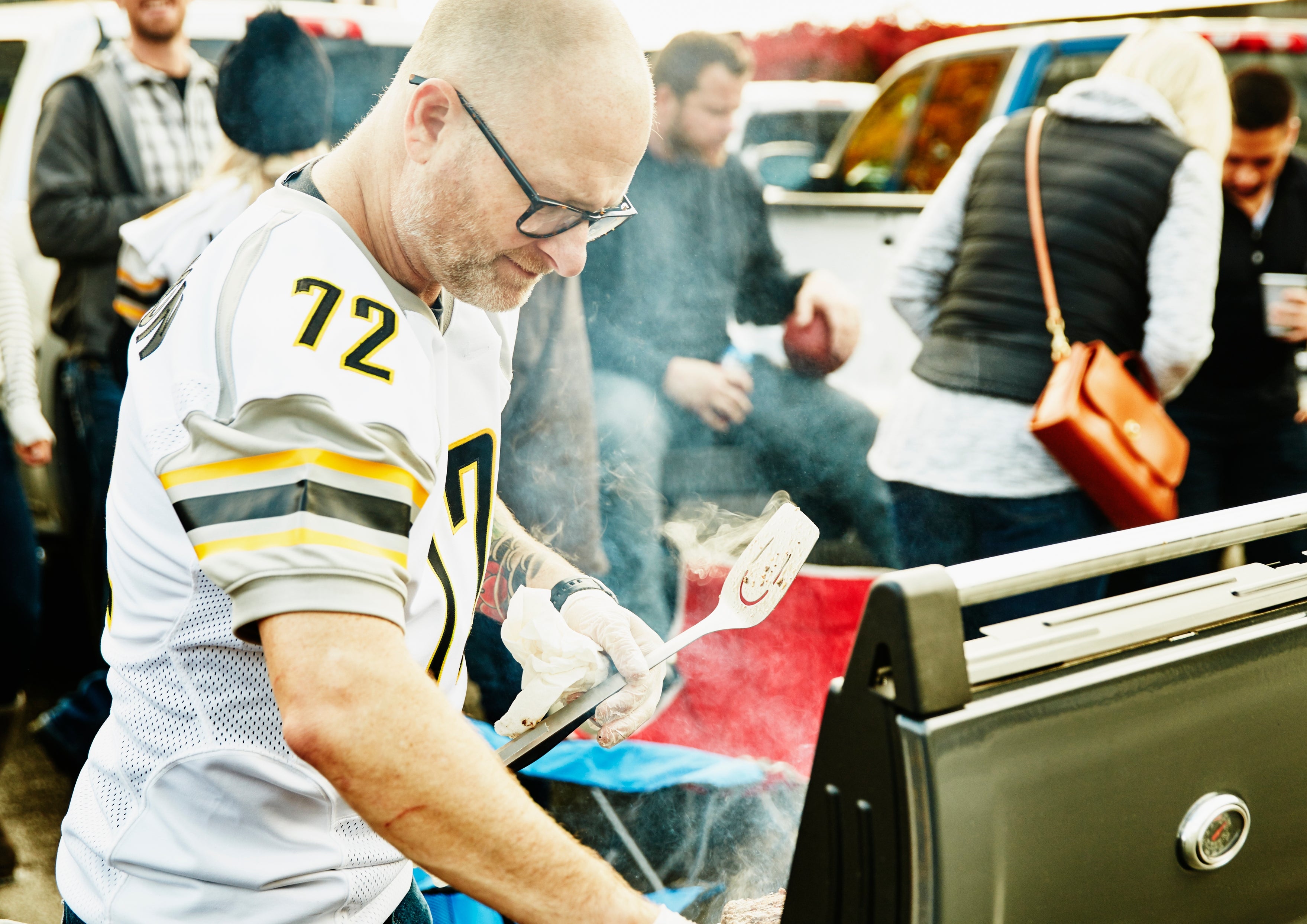 Person grilling nutrient  astatine  a tailgate party, surrounded by radical   successful  sports jerseys and jackets, adjacent   parked cars