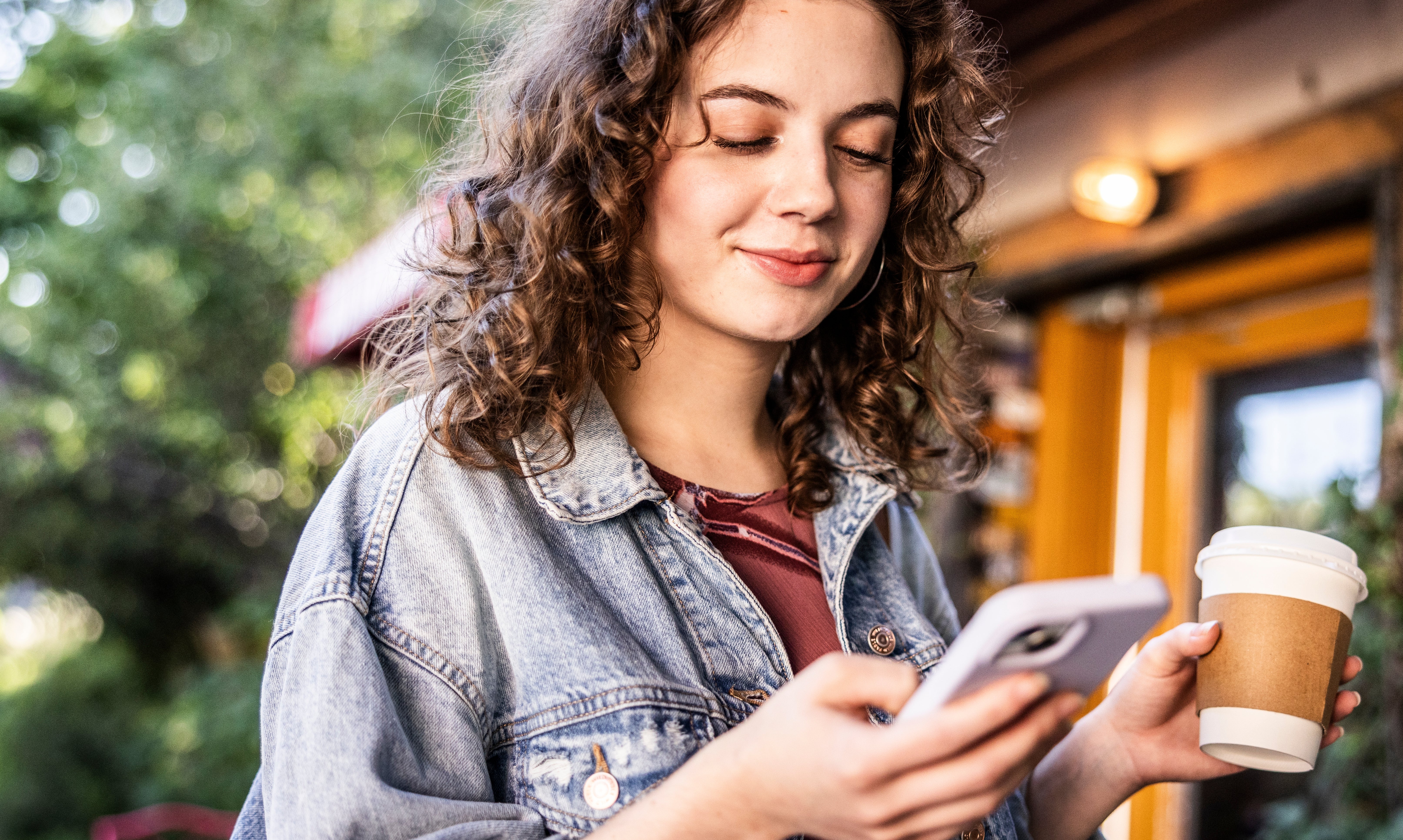 Person with curly hairsbreadth  smiles, holding a smartphone and java  cup, lasting  outdoors successful  a casual setting