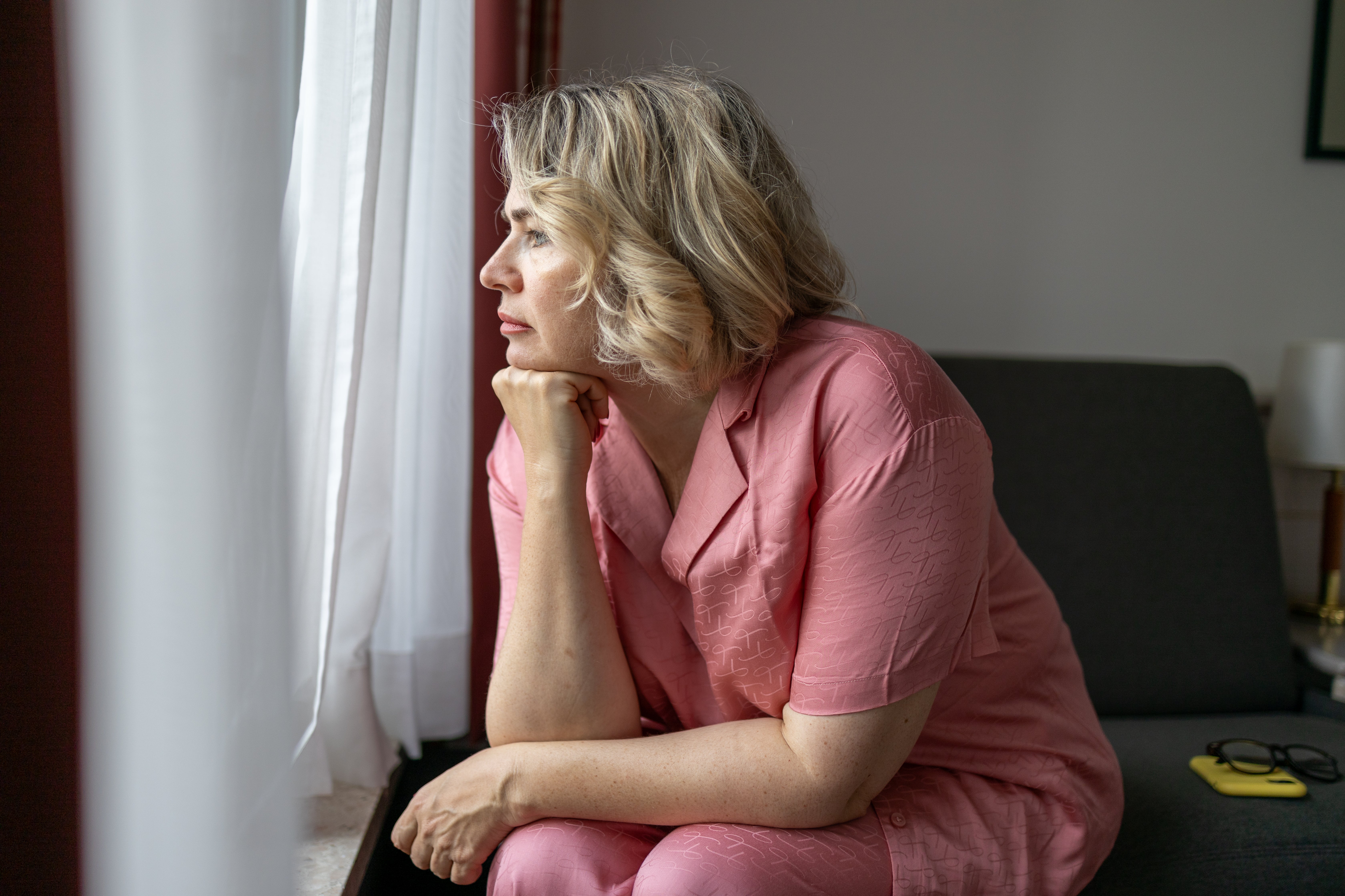 Person successful  casual attire gazes thoughtfully retired  a window, seated connected  a sofa with eyeglasses and a telephone  nearby