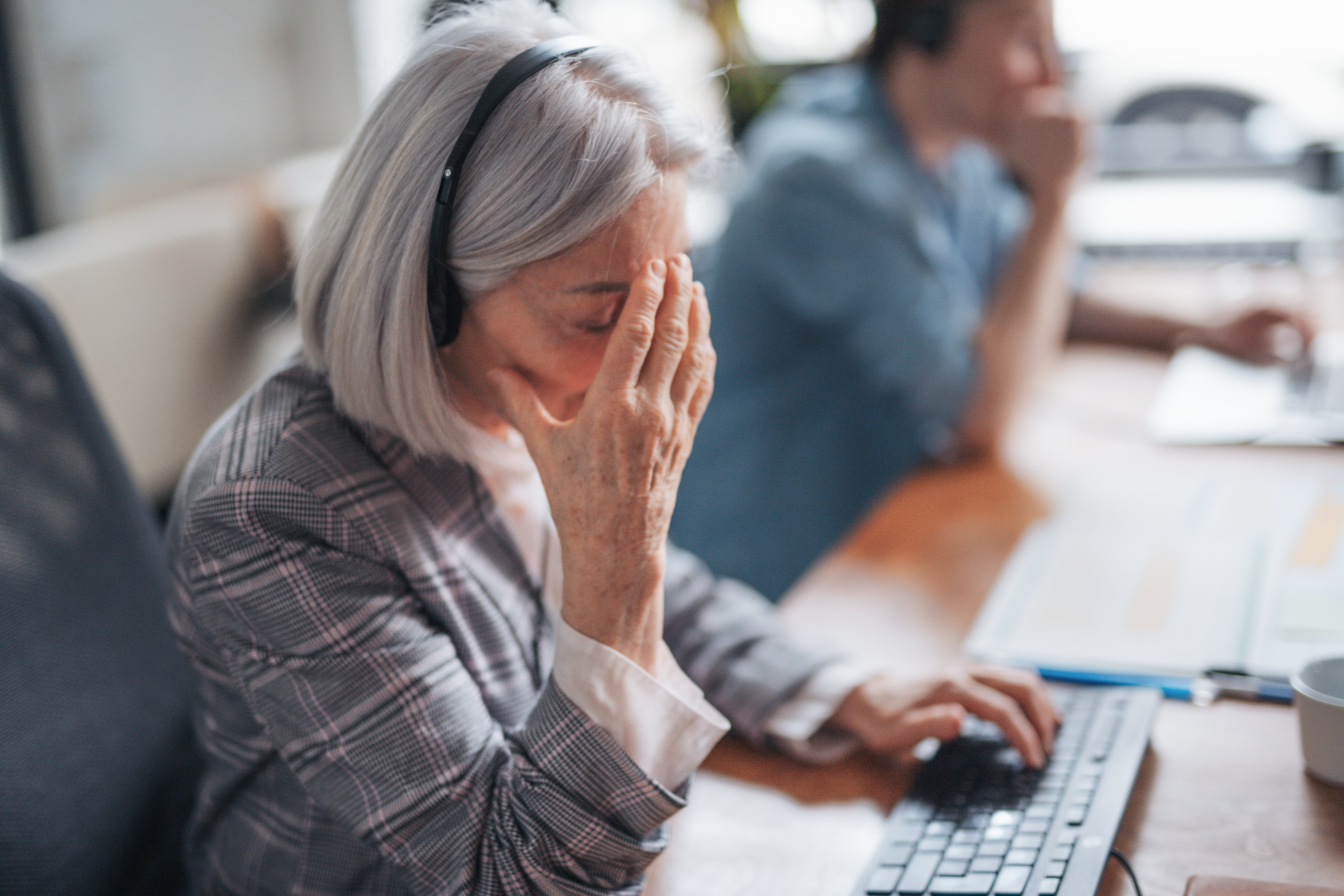 A idiosyncratic   with headphones astatine  a table  looks stressed, with their manus  connected  their forehead, portion    utilizing a computer