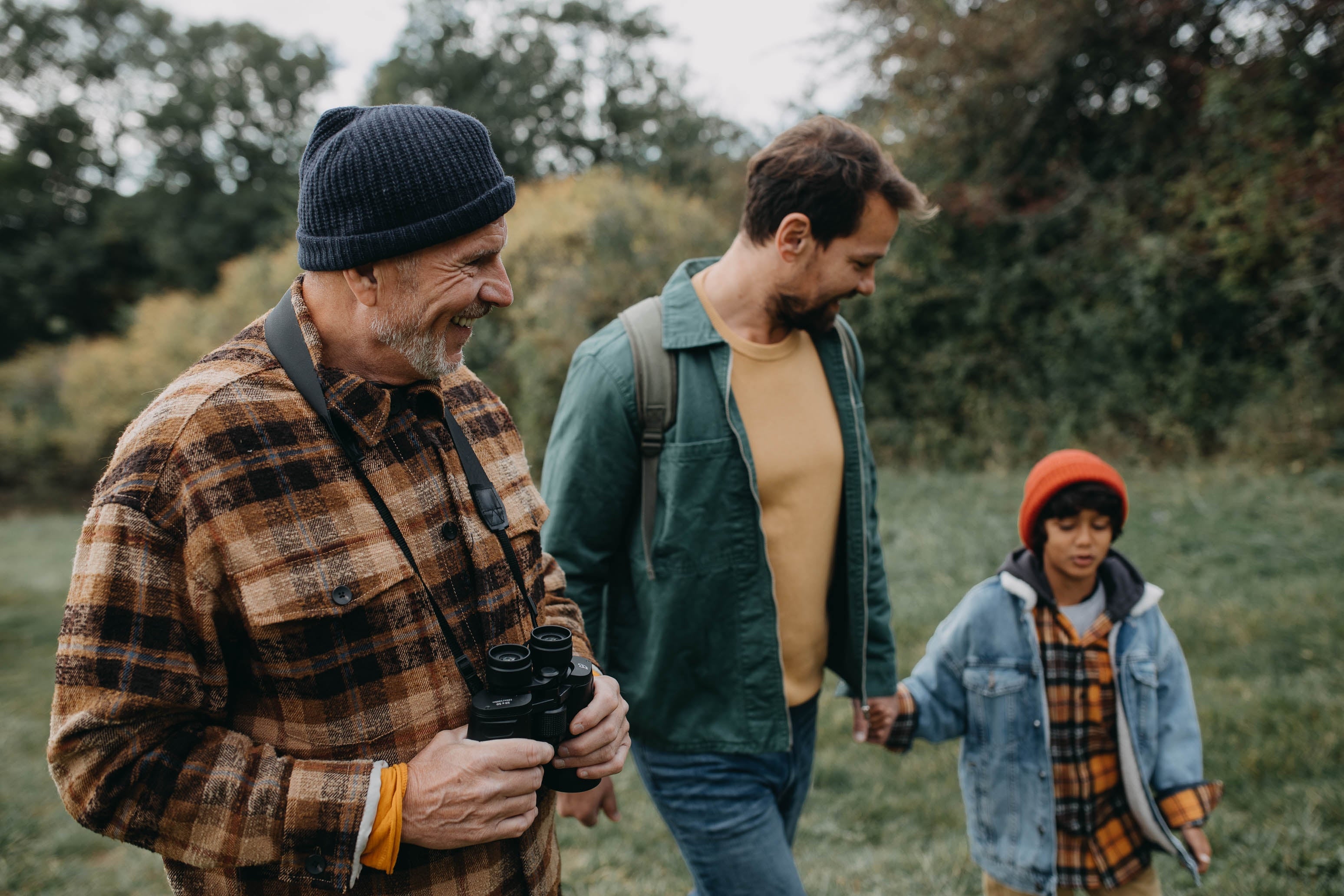  an older antheral   with binoculars, a younger man, and a kid  successful  a beanie. They look  to beryllium  enjoying a quality  walk